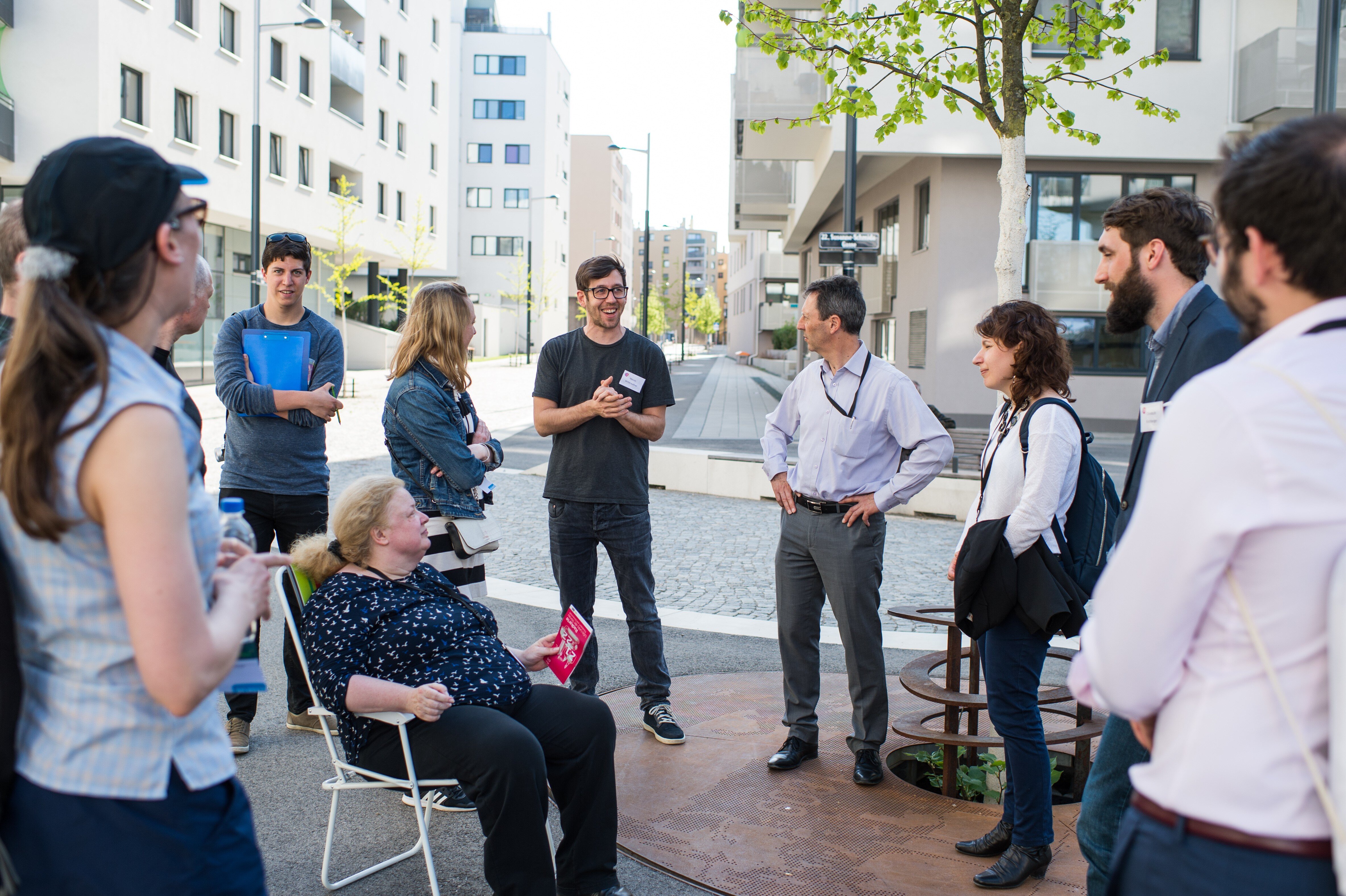 Mehrere Personen stehen im Öffentlichen Raum, eine Frau sitzt auf einem Campingstuhl, ein Mann spricht, die anderen sehen ihn an, im Hintergrund mehrere Häuser