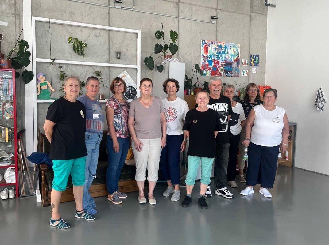 Zehn Personen stehen nebeneinander in einem Raum und blicken in die Kamera, im Hintergrund sind Bilder und Pflanzen an der Wand