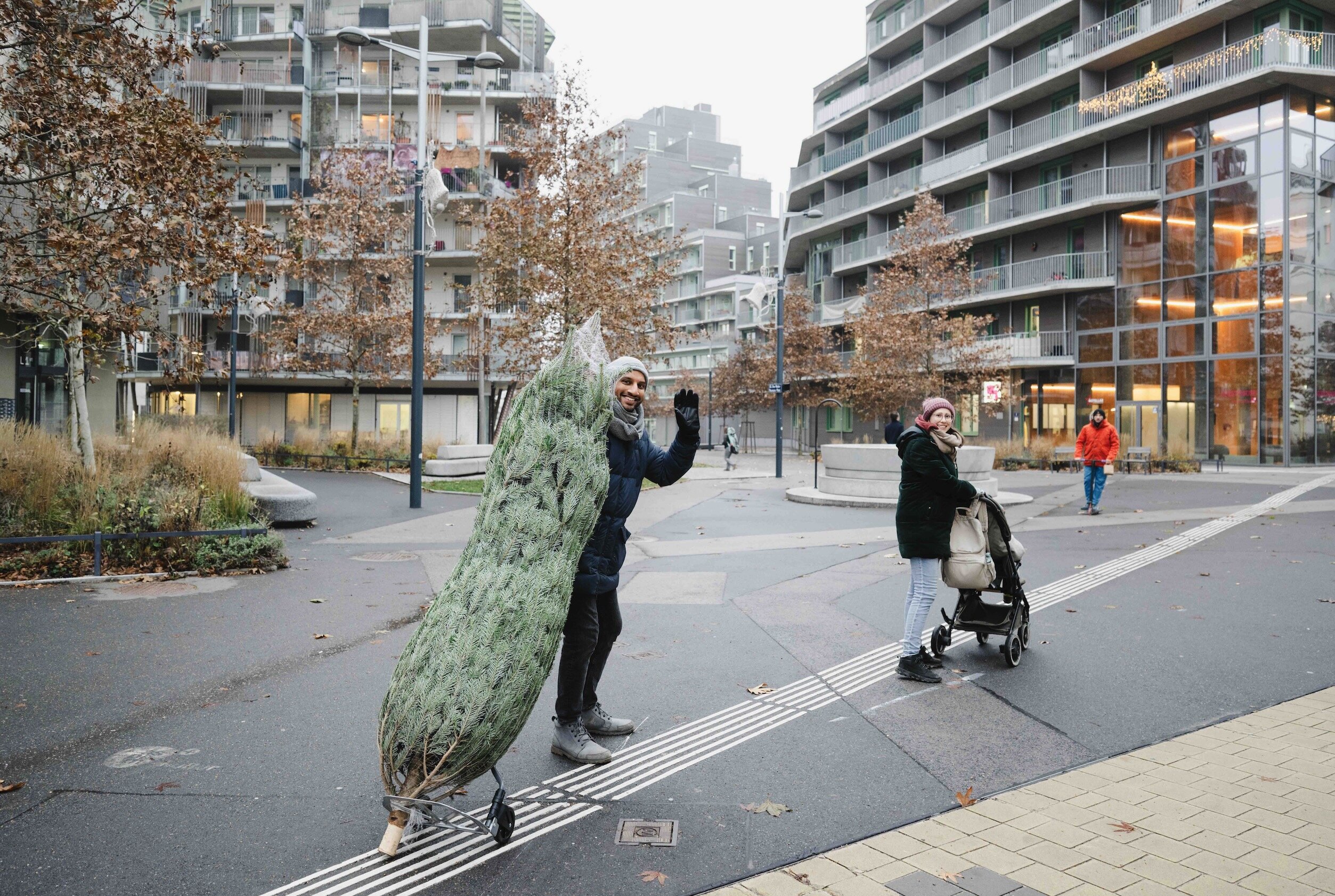 Mann winkt in die Kamera und transportiert einen Christbaum in der Straße, etwas weiter hinten steht eine Frau mit Kinderwagen und blickt ebenfalls in die Kamera