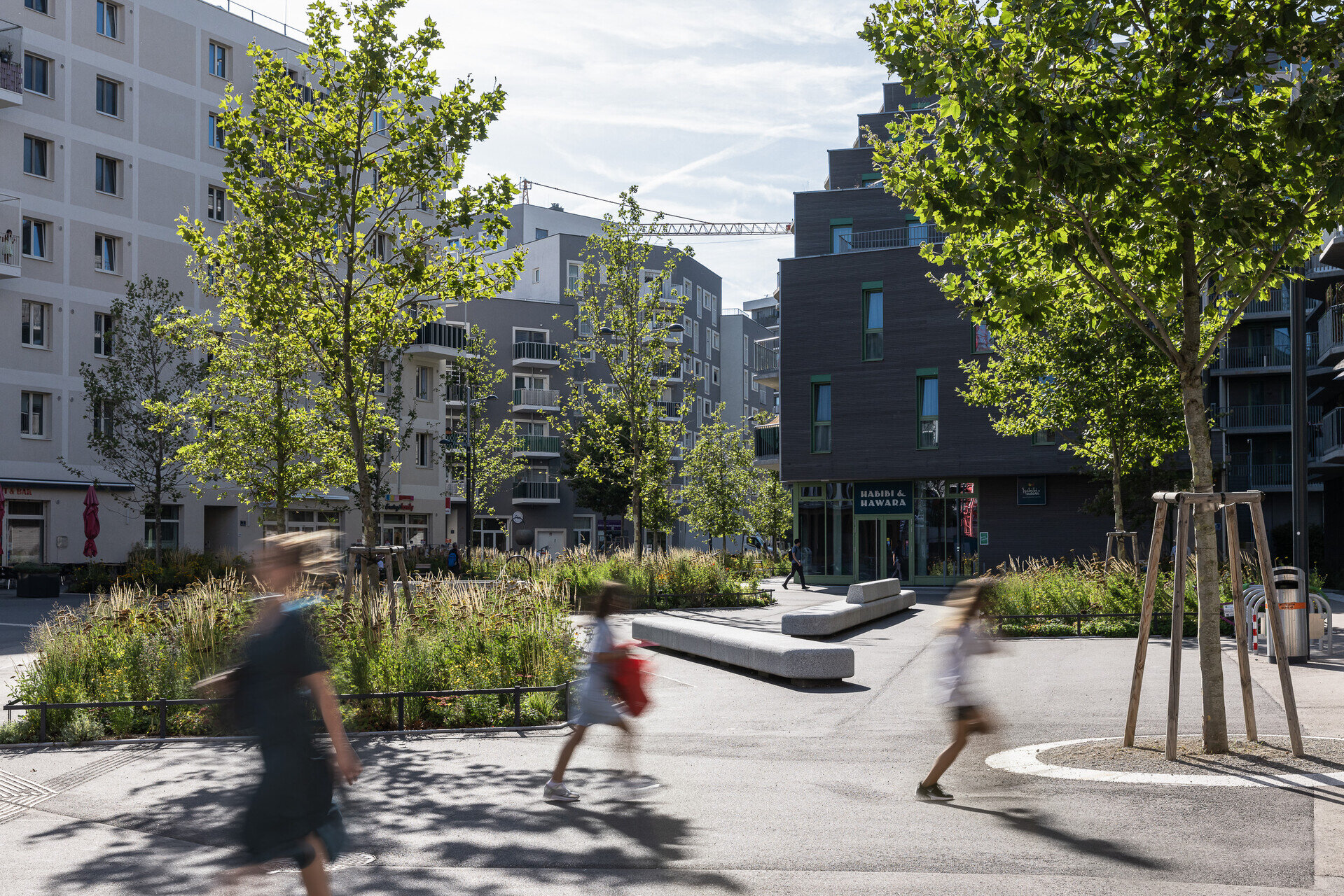 Platz in der Seestadt mit Bäumen und Passanten, die durch das Bild gehen.