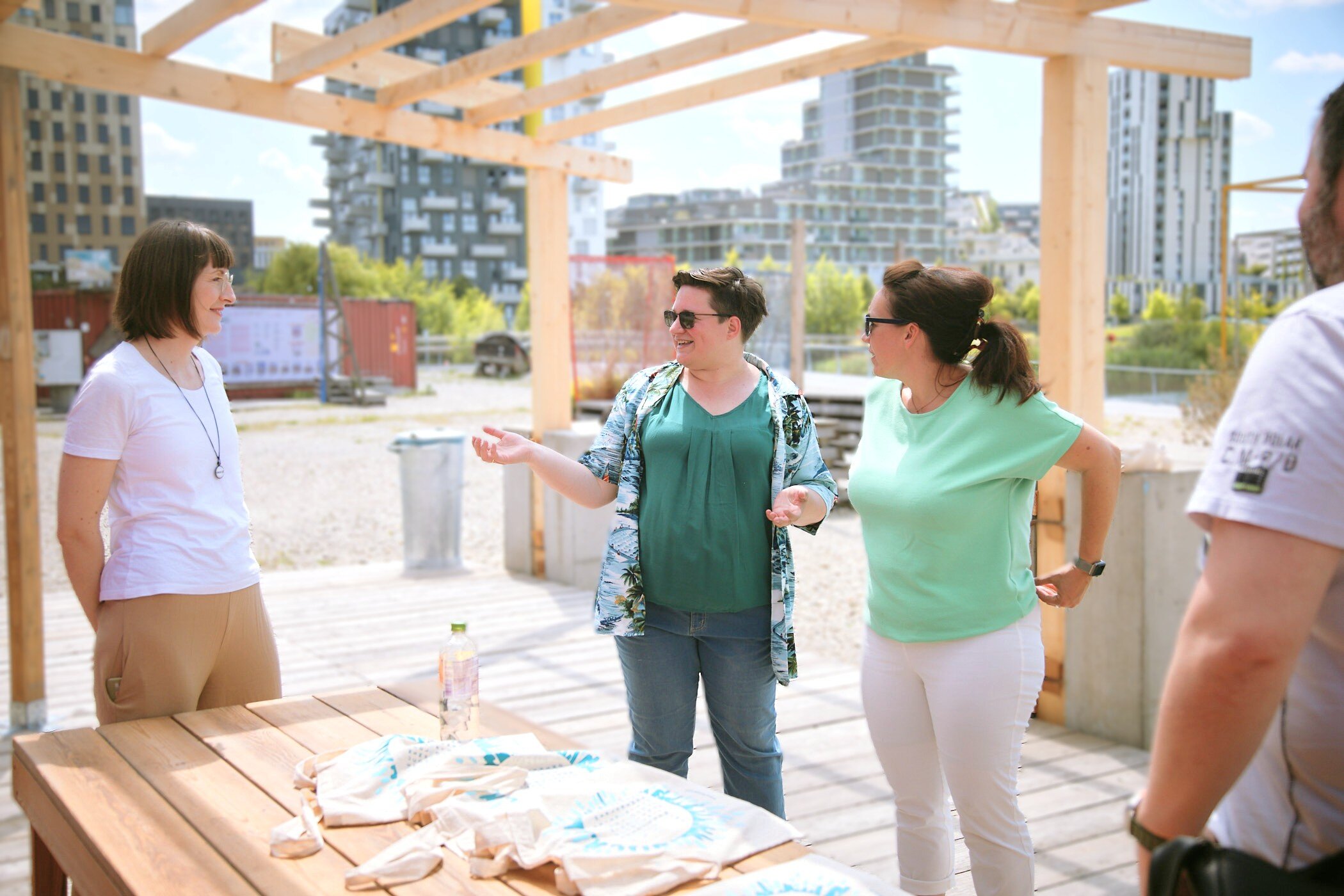 drei Frauen stehen zusammen unter einer Holzkonstruktion, unterhalten sich, auf dem Tisch vor ihnen liegen Stoffsackerl