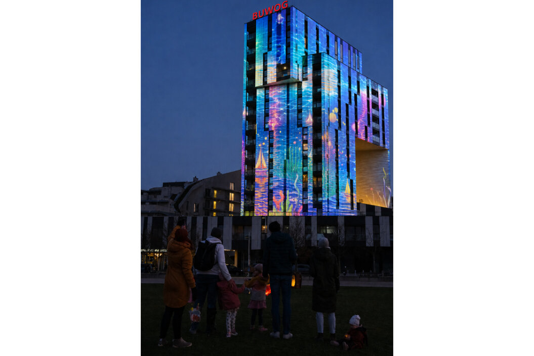 Menschen stehen vor einem Gebäude in der Wiese, es ist dunkel, die Fassade des Gebäudes ist bunt beleuchtet durch eine Lichtprojektion und alle schauen sich die Projektion an 