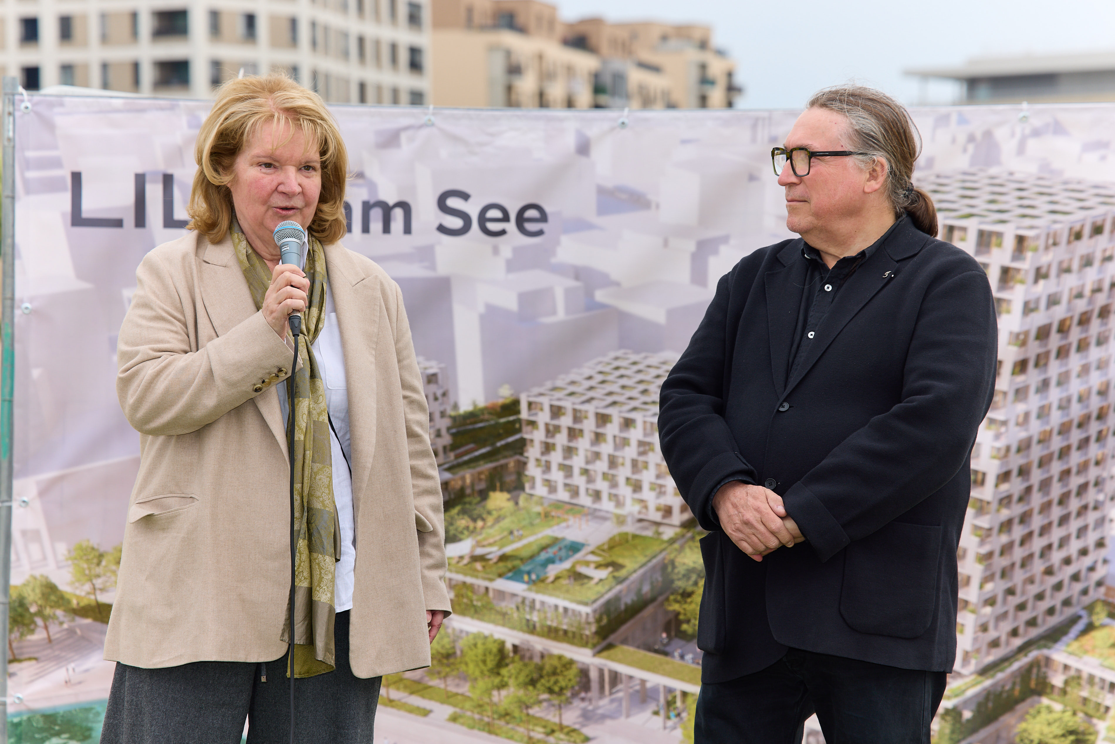 Frau spricht bei Baustart-Event von LILY am See, Mann steht daneben vor Projektbanner.