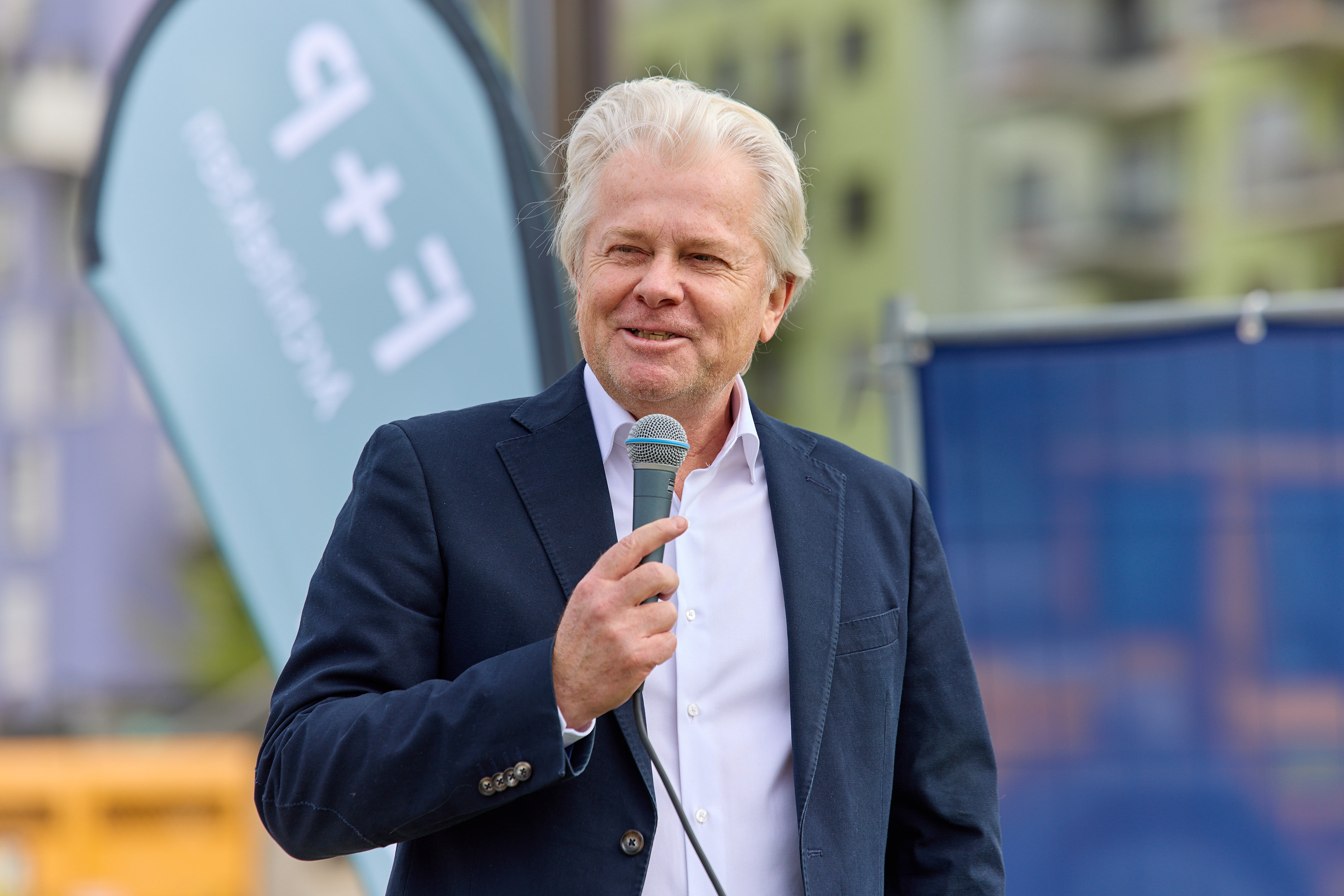 Mann im Anzug hält Rede mit Mikrofon bei Spatenstich-Veranstaltung des Bauprojekts LILY am See.