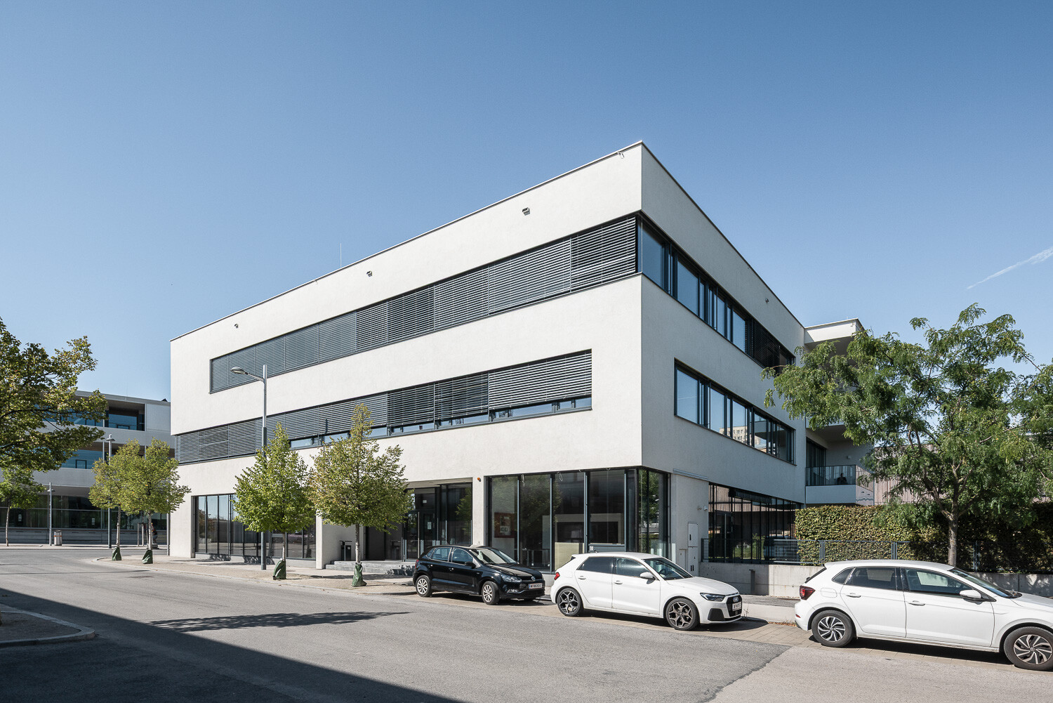 Modernen Bürogebäudes mit weißer Fassade und großen, rechteckigen Fenstern. Parkende Autos stehen davor.