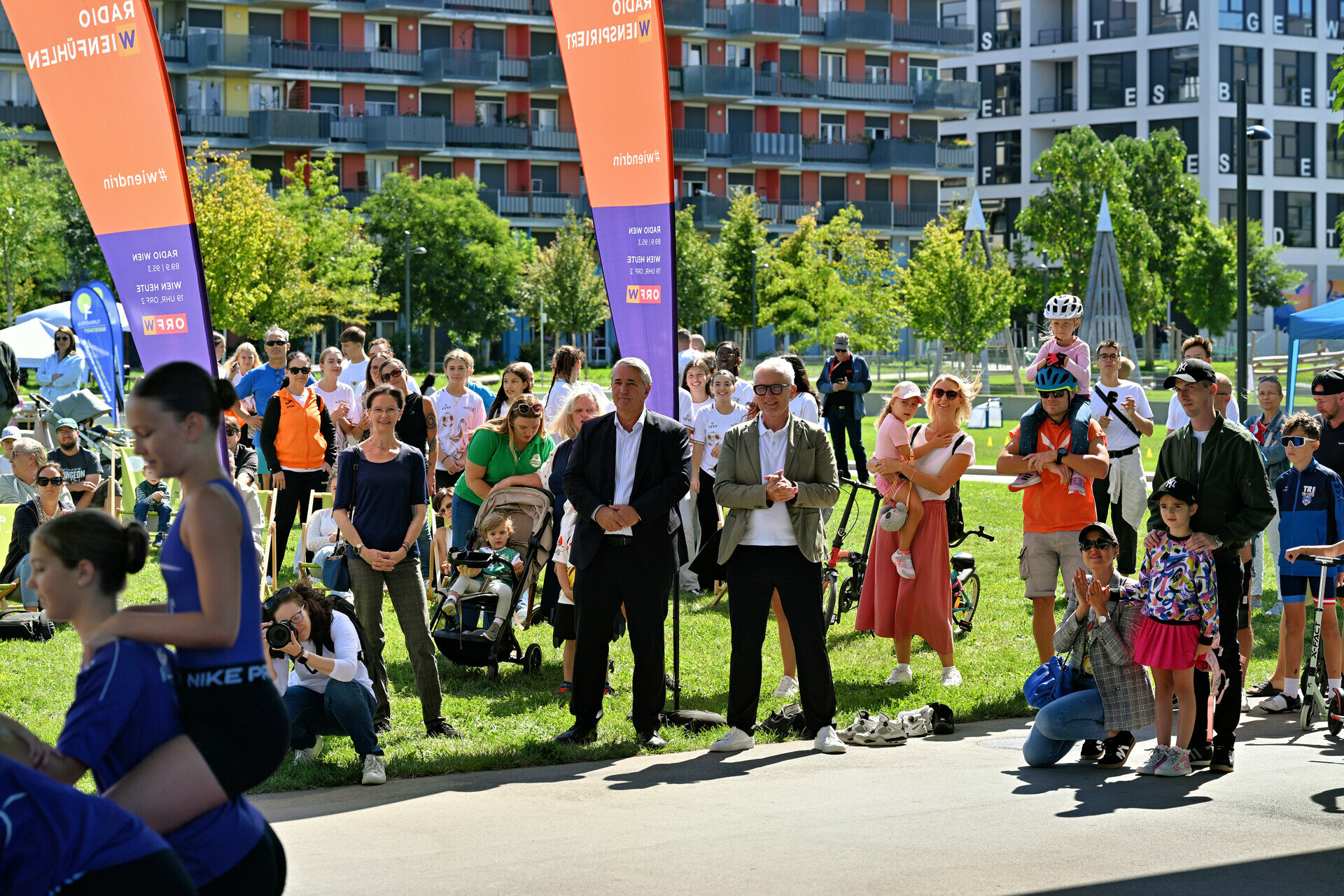 Bezirksvorsteher Ernst Nevrivy und Wien 3420-Vorstand Robert Grüneis bei der Eröffnung des 1. Sportfests Donaustadt. Mehrere Personen auf einer grünen Wiese vor Gebäuden.