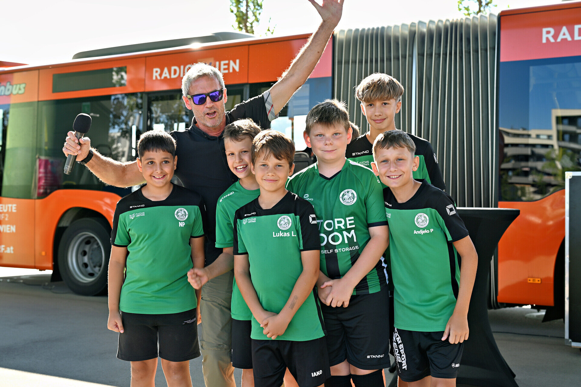 Fußballnachwuchs des FC Hellas Kagran mit Tormannlegende Michael Konsel. Moderator mit Kindern in Sportdressen vor einem orangen Bus.