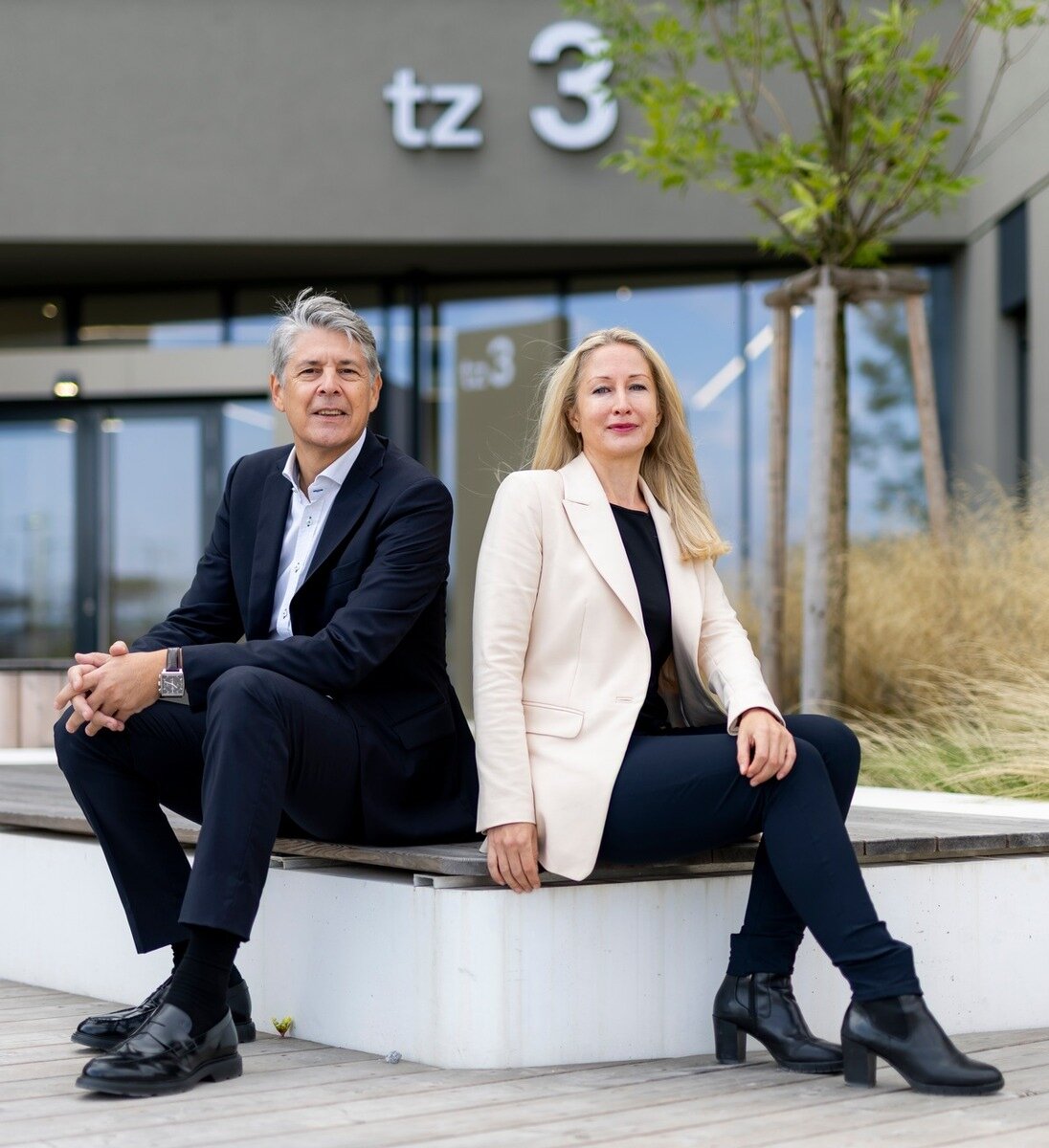 Zwei Personen in Businesskleidung sitzen vor dem Gebäude „tz3“ des Technologiezentrums Seestadt auf einer Holzbank. Beide blicken in die Kamera; im Hintergrund sind Glasfassade und junge Bepflanzung zu sehen.