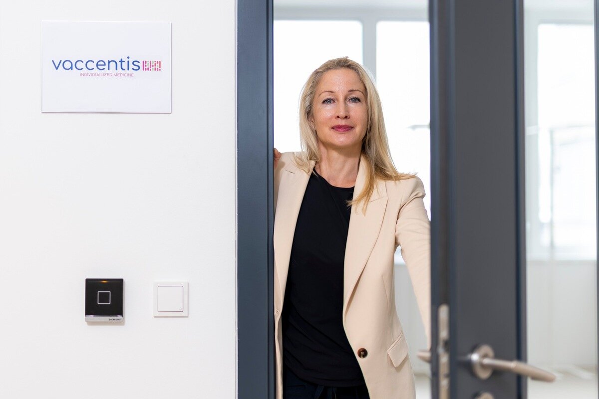 Eine Person in heller Blazerjacke steht in einer halb geöffneten Tür eines Büroraums. An der Wand hängt ein Schild mit dem Vaccentis-Logo; im Hintergrund ist ein heller Raum mit Fenstern sichtbar.