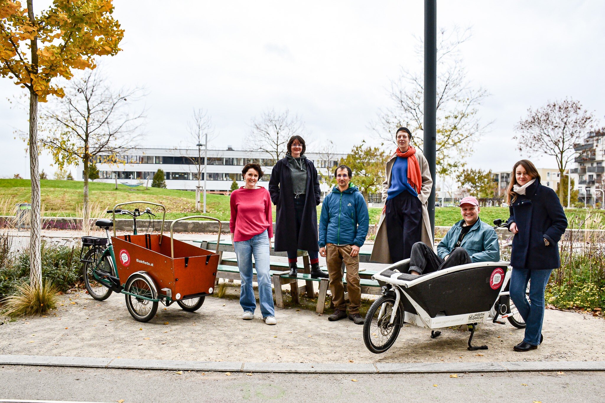 Sechs Personen stehen auf Schotte und blicken in die Kamera, eine Person sitzt in einem Lasten Lastenrad, im Hintergrund Wiese, Bäume und ein Gebäude