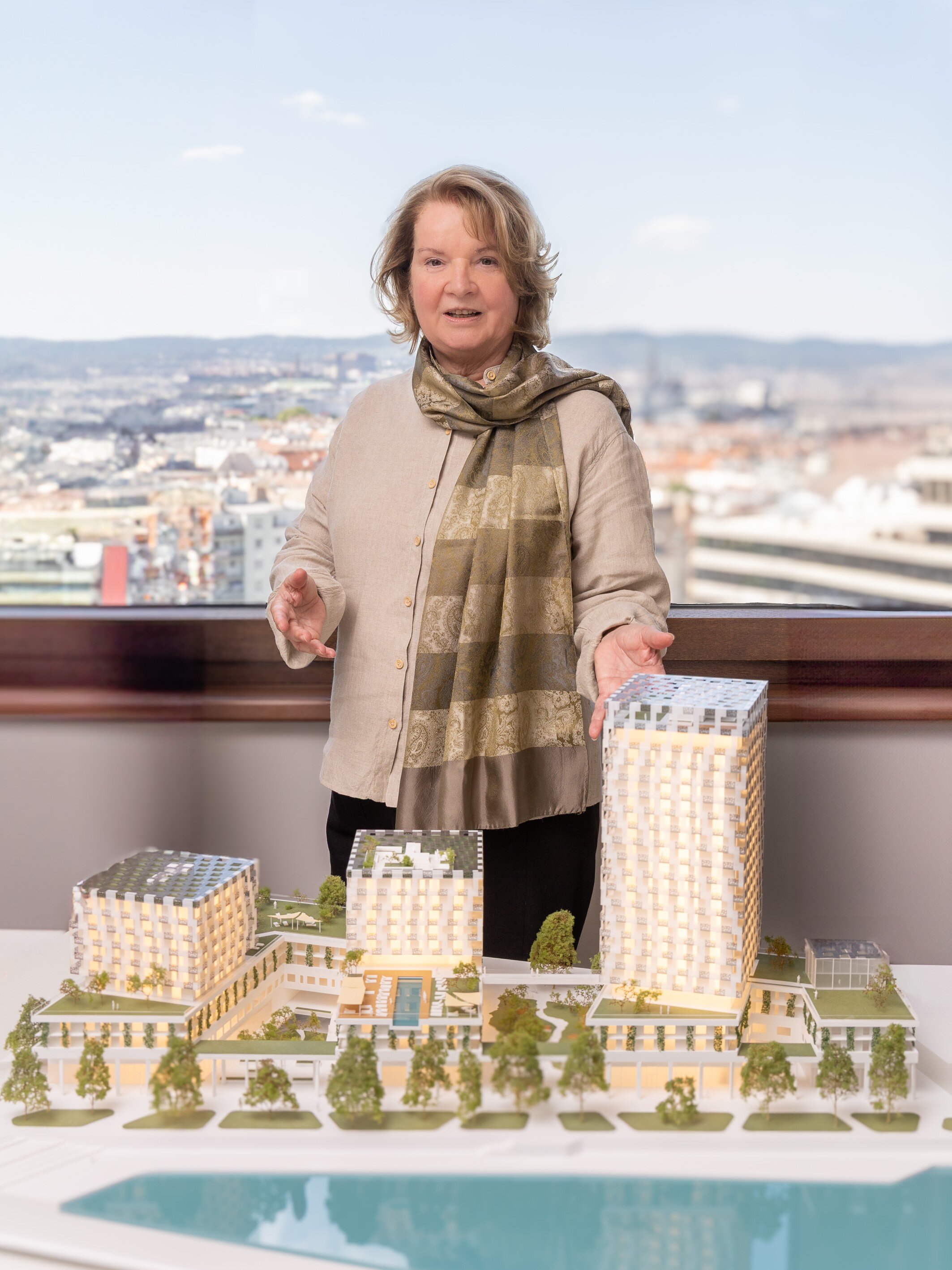 Blonde Frau steht vor einem Architekturmodell mit Hochhäusern und Grünflächen. 