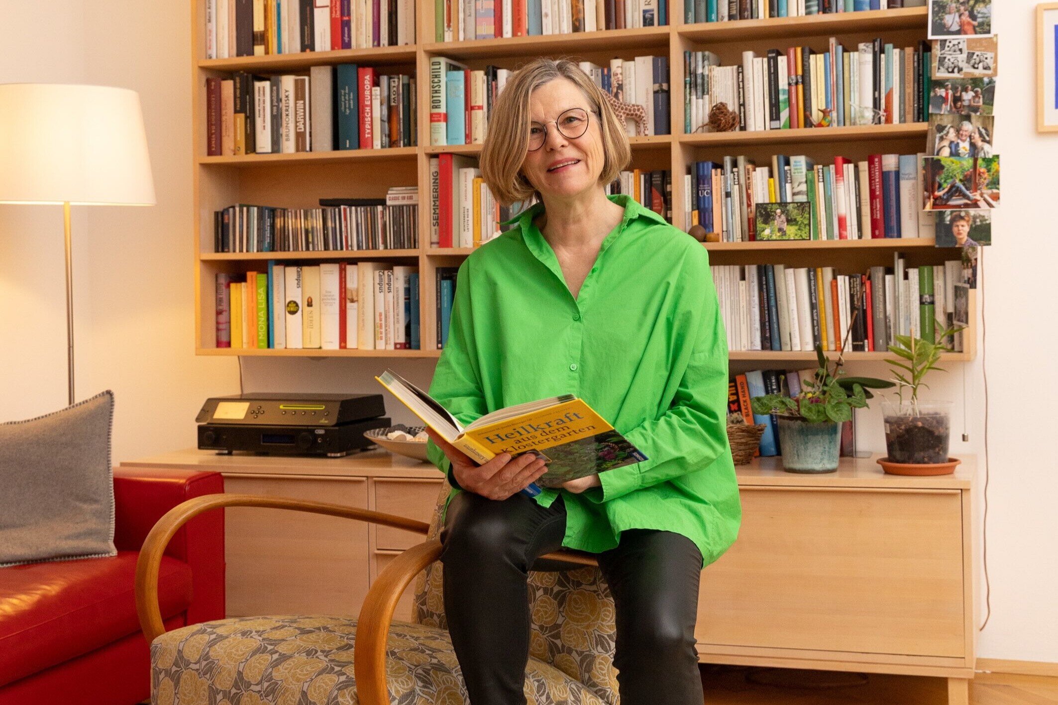 Frau sitzt vor einem Bücherregal und hält ein Buch in der Hand. 