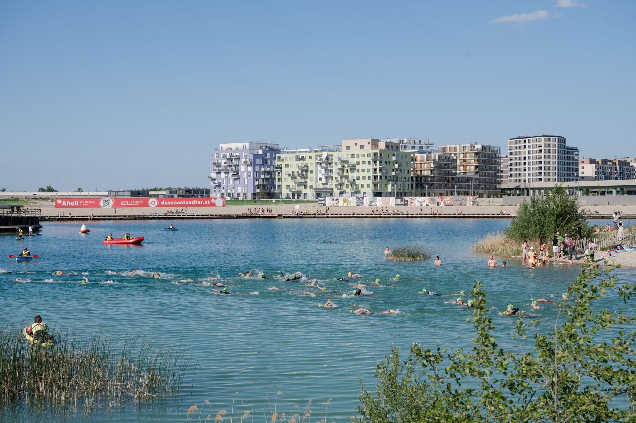 See, wo viele Schwimmer sich im Wasser tummeln. Im Hintergrund Hochhäuser.