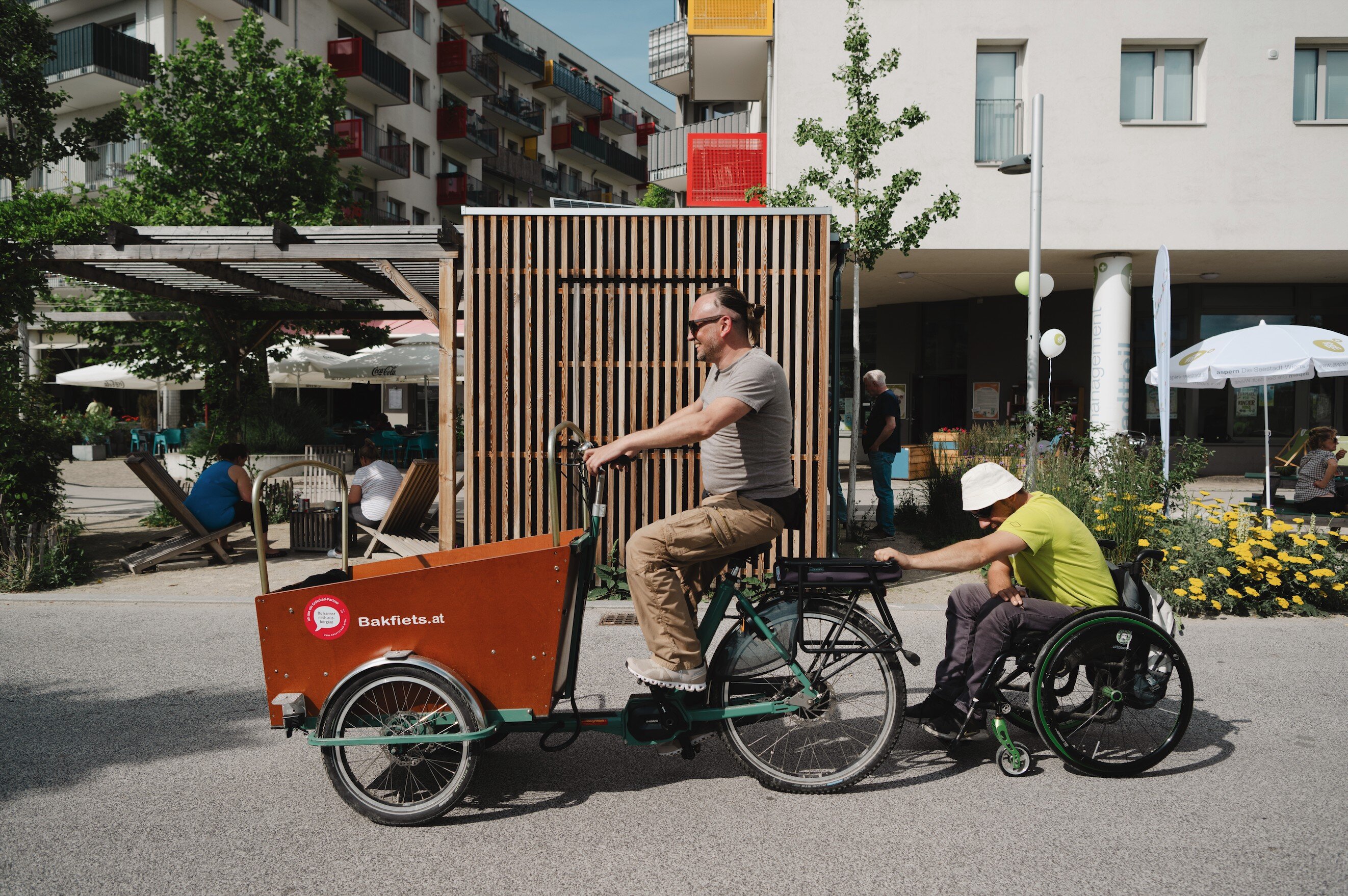 Ein Rollstuhlfahrer, der ein Lastenrad mit einer Person drauf anschiebt. 