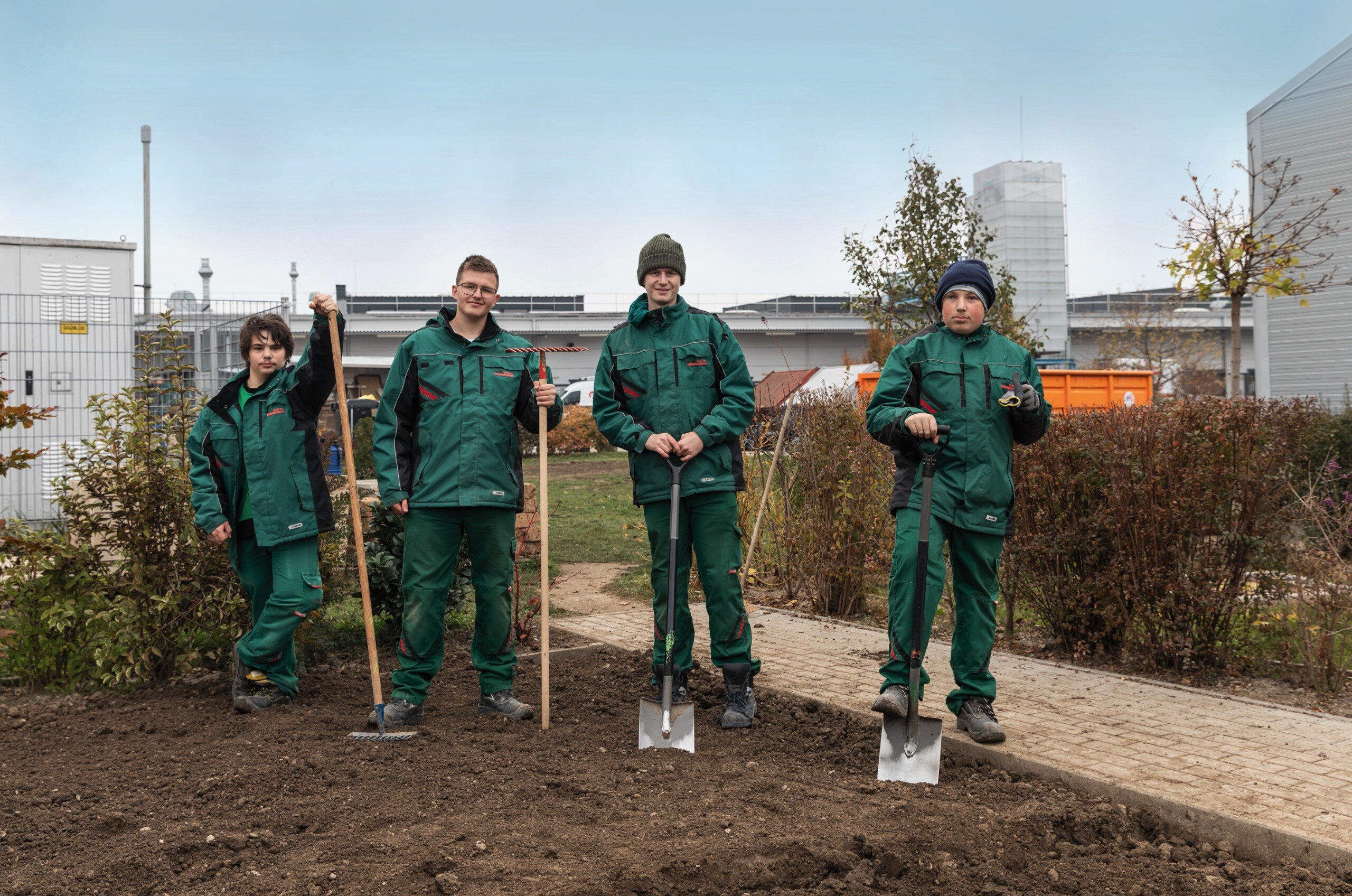 Vier Männer in grüner Arbeitsbekleidung und Gartengeräten stehen auf einem Boden aus Erde.