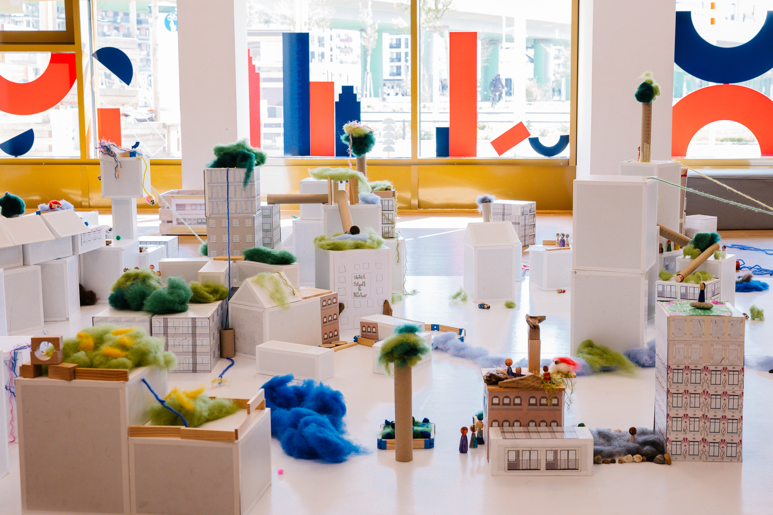 Ein großes Stadtmodell, gebaut aus Kartons, Papier, Papierrollen und Filz, das über den gesamten Raum verteilt auf dem Boden steht. 