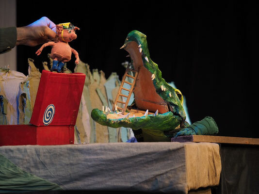 Theaterbühne eines Puppentheaters, ein Schwein mit einem Zahnputzkoffer untersucht die Zähne eines Krokodils mit weit aufgesperrtem Maul 