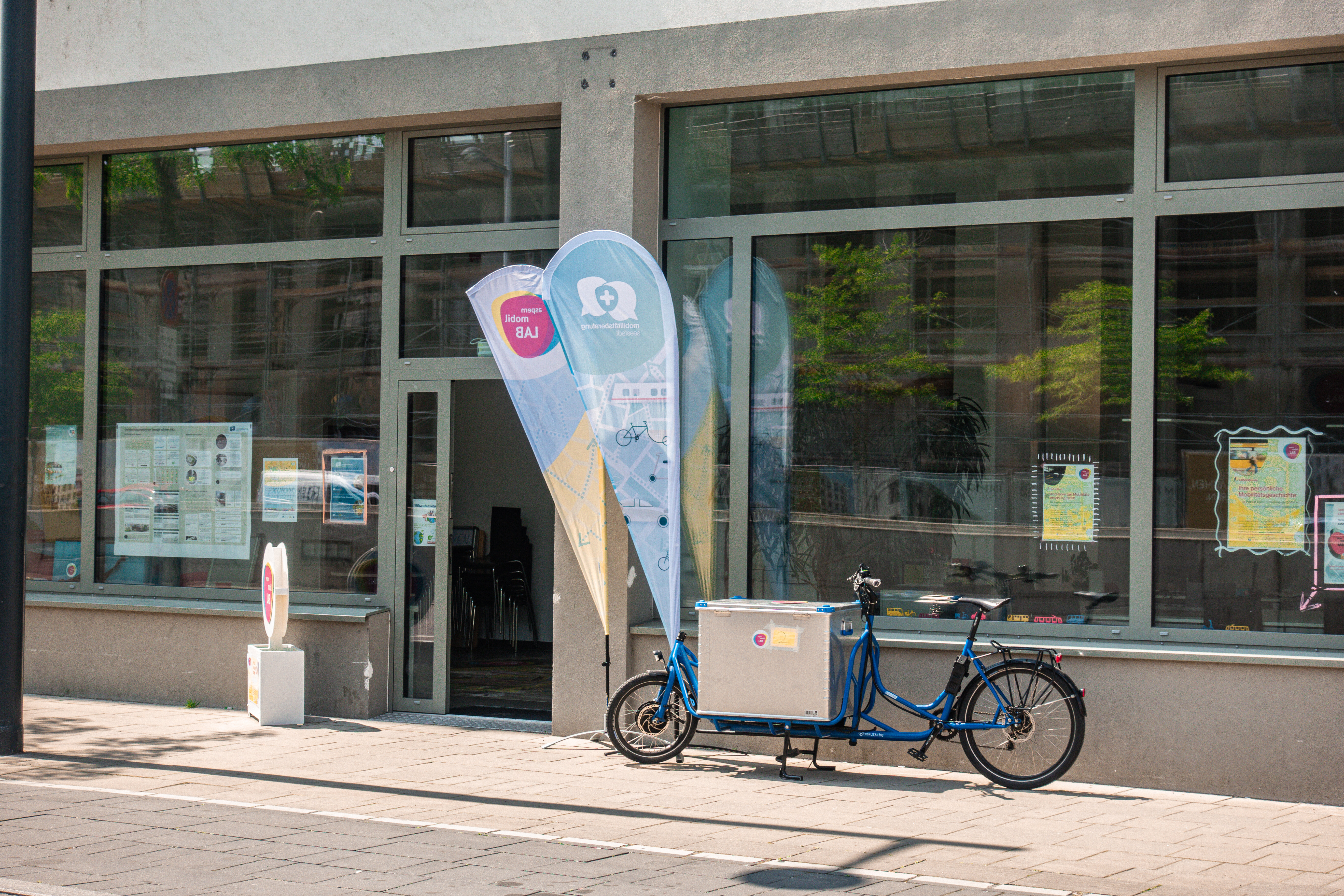 Ein blaues Lastenrad mit Container vorne steht vor einer Glasfront eines Gebäudes mit Türe. 
