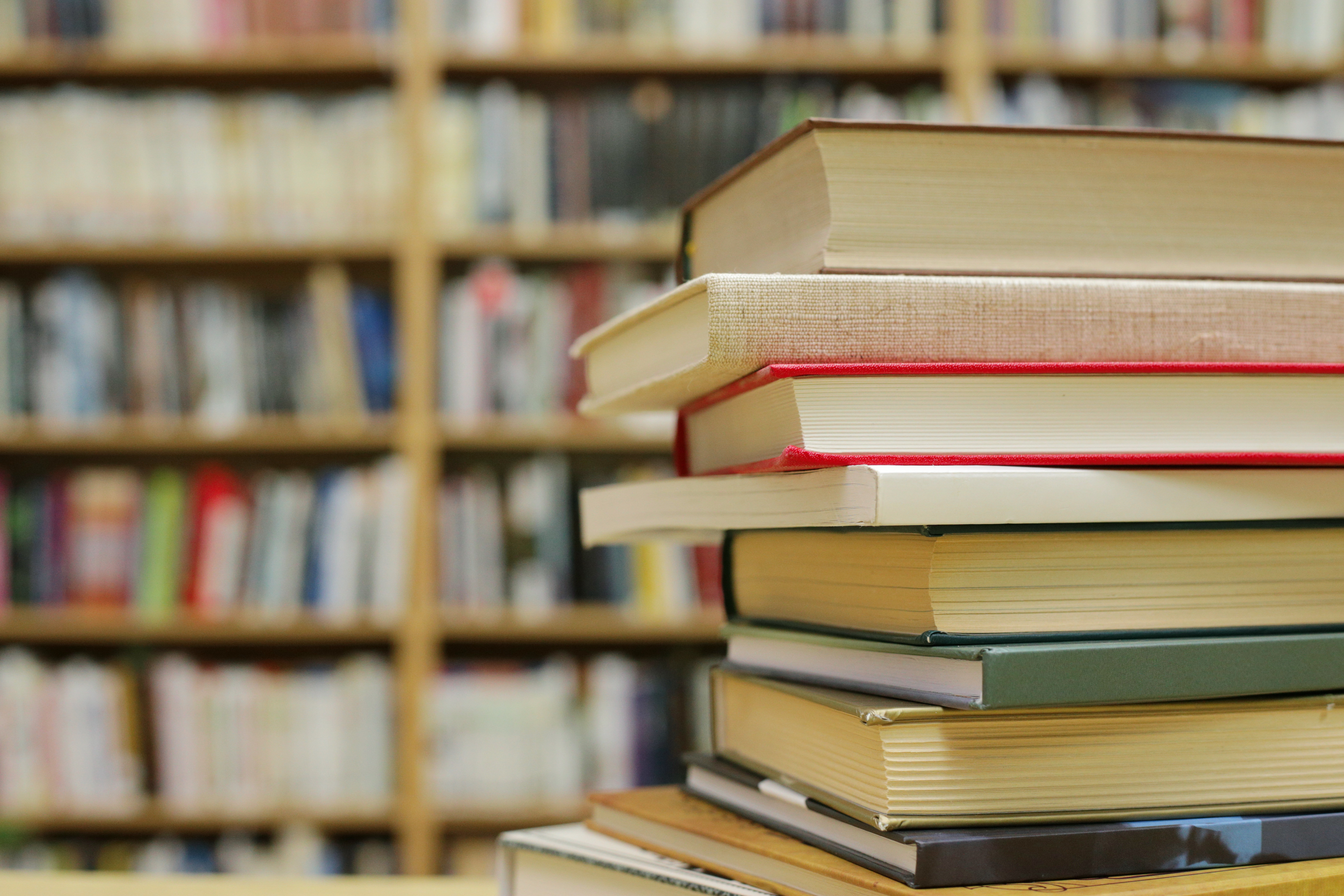 Bücherstapel mit 10 unterschiedlich dicken Büchern, die aufeinander liegen und im Hintergrund ist ein verschwommenes Bücherregal zu sehen