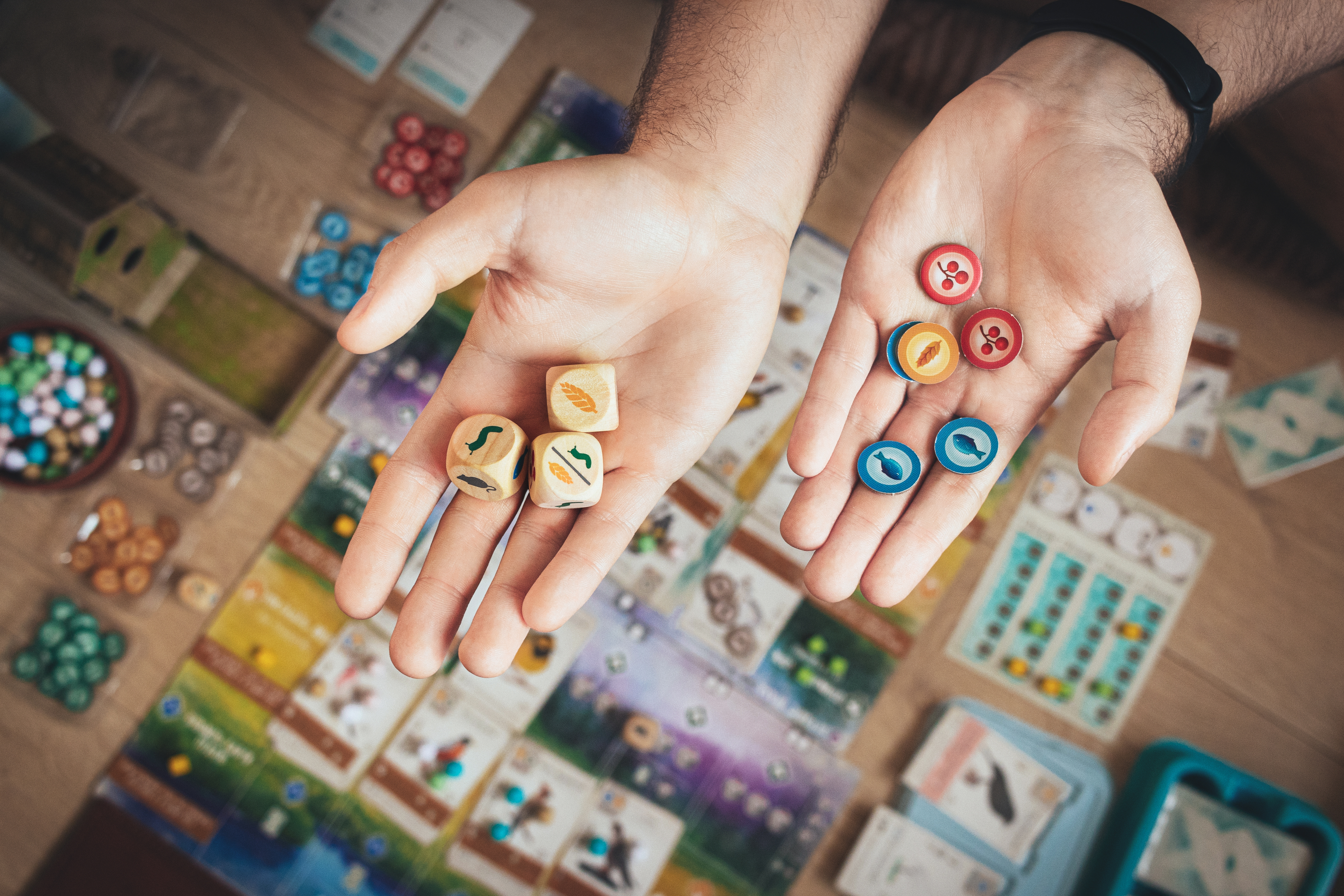 Zwei Hände mit nach oben offen ausgerichteter Handfläche, in der linken Hand sind 3 Spielwürfel, in der rechten Hand Spielchips in verschiedenen Farben zu sehen. Im Hintergrund ist ein Tisch mit Karten und Brettspielen zu erkennen.