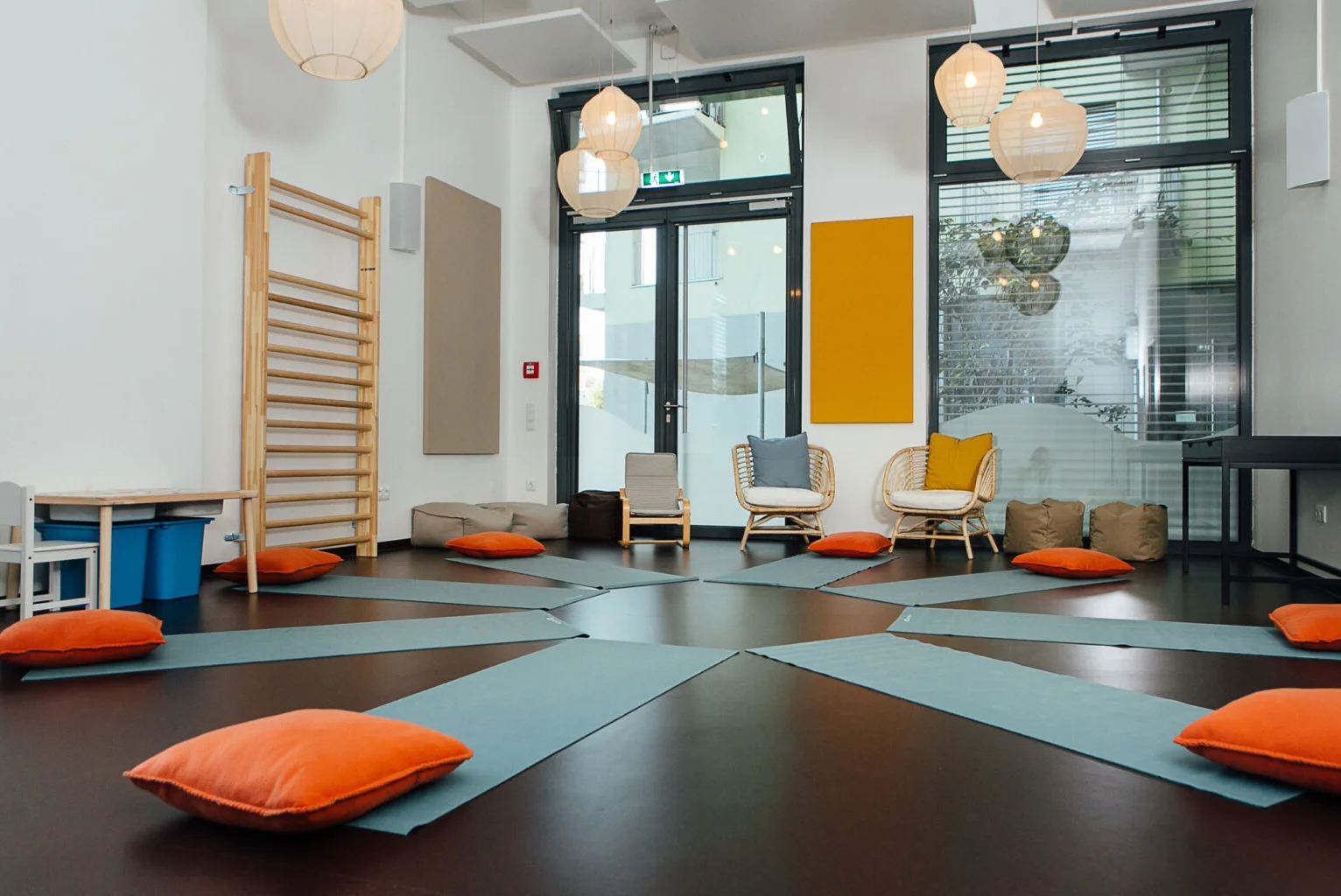 Hellblaue Yogamatten liegen ringförmig aufgelegt mit einem orangefarbenem Polster am Kopfende in einem kleinen Sportraum, im Hintergrund sind Fenster mit einem Blick in den Innenhof