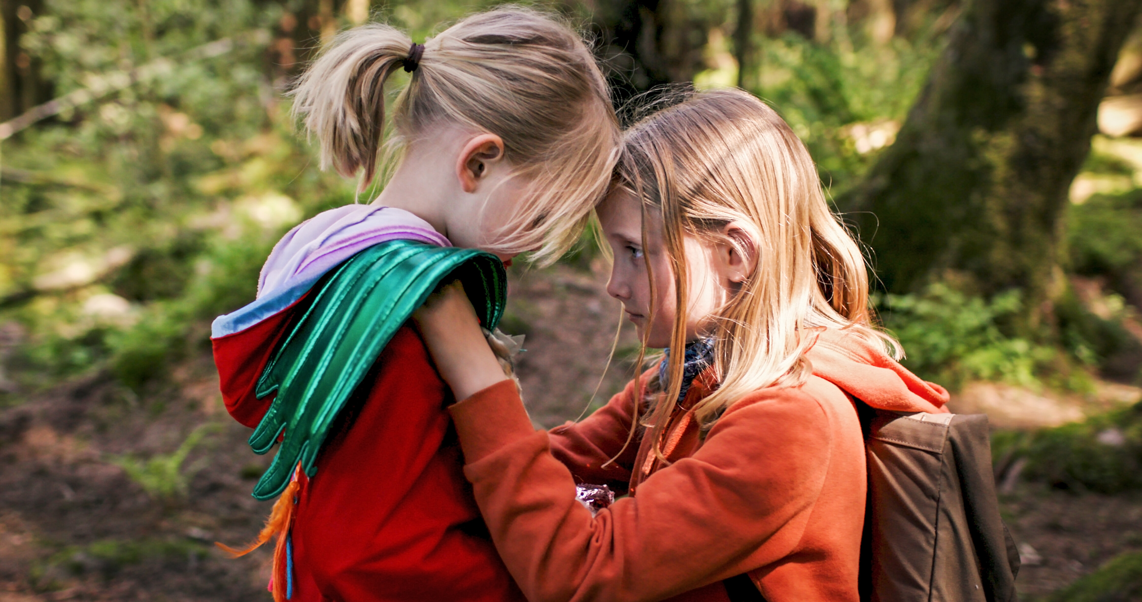Ein blondhaariges Mädchen in einem orangefarbenem Pullover kniet vor einem anderen blonden Mädchen mit orangefarbenen Pullover und sie haben die Köpfe andeinandergelegt, sie stehen im Wald