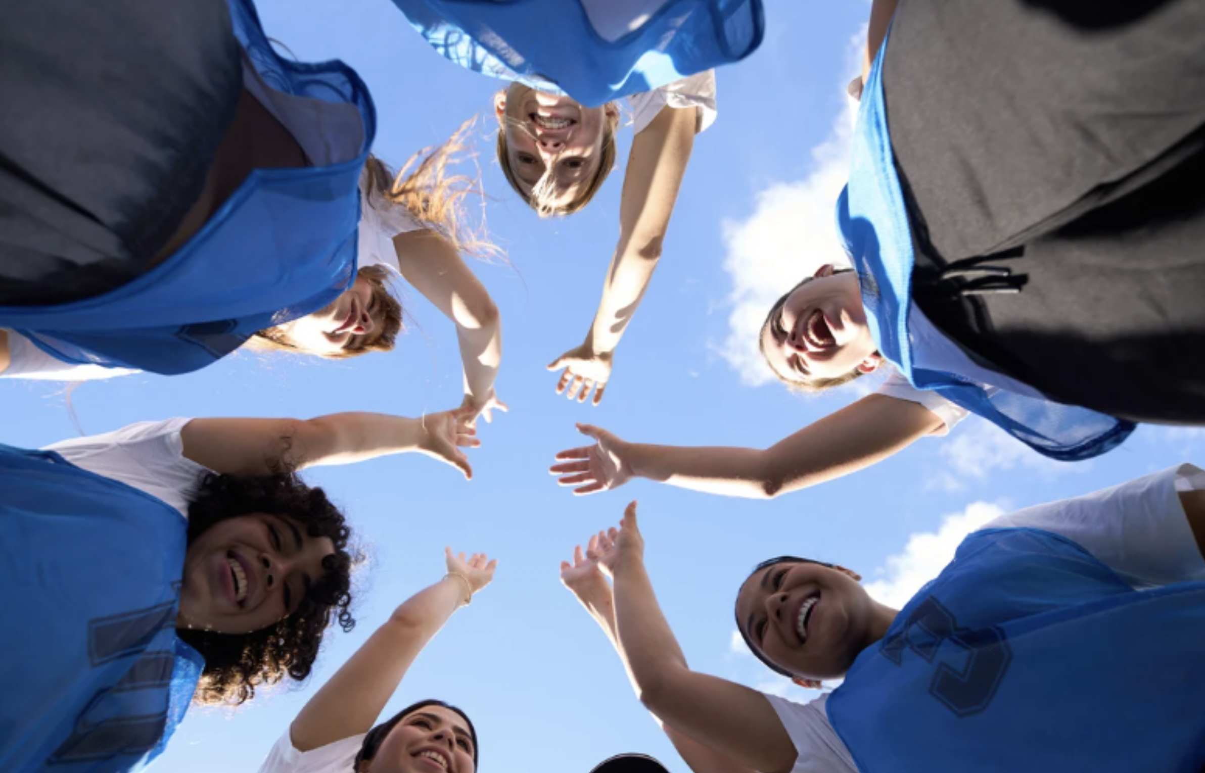 Gruppe von 7 sportlich gekleideten Mädchen stehen im Kreis und strecken in der Mitte die Hände zusammen, das Foto wurde von der Mitte des Kreises aufgenommen und zeigt die Mädchen von unten