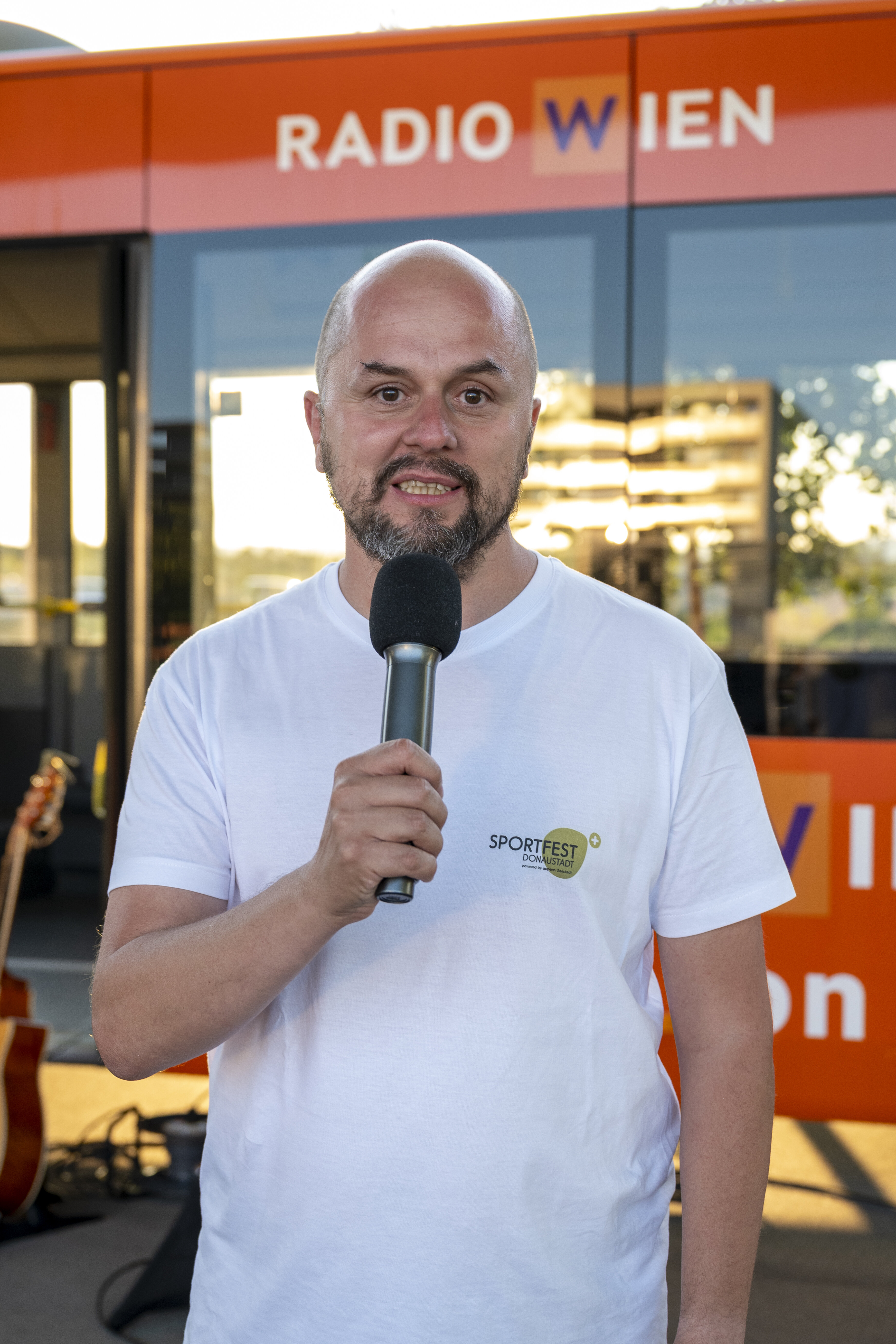 Mann mit Mikrofon steht auf einer Bühne, im Hintergrund das Logo von Radio Wien. Auf seinem weißen Leiberl steht Sportfest Donaustadt