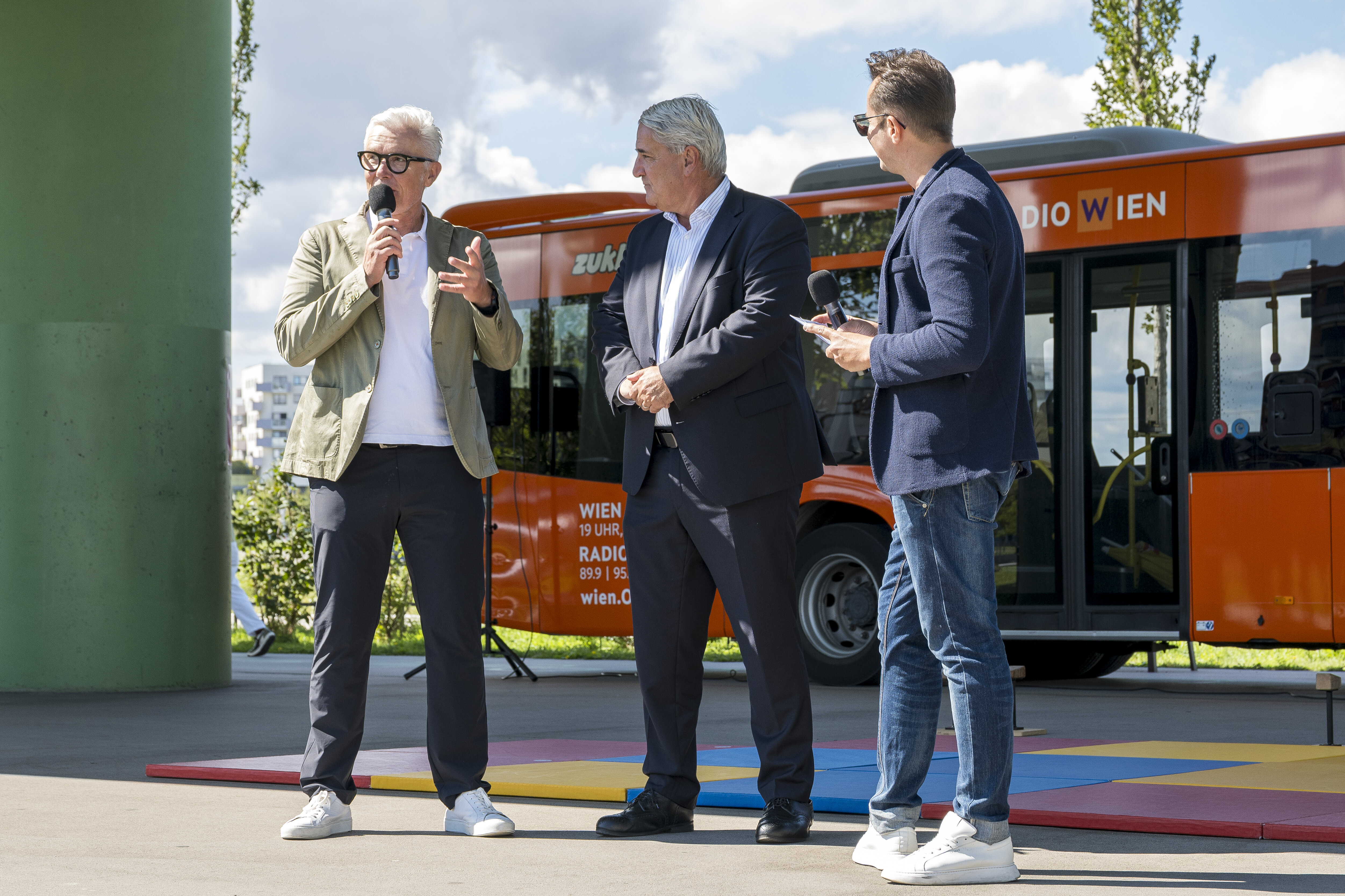 Geschäftsführer Robert Grüneis und Bezirksvorsteher Ernst Nevrivy stehen mit einem Moderator unter der U-Bahntrasse und sprechen in ein Mikrofon