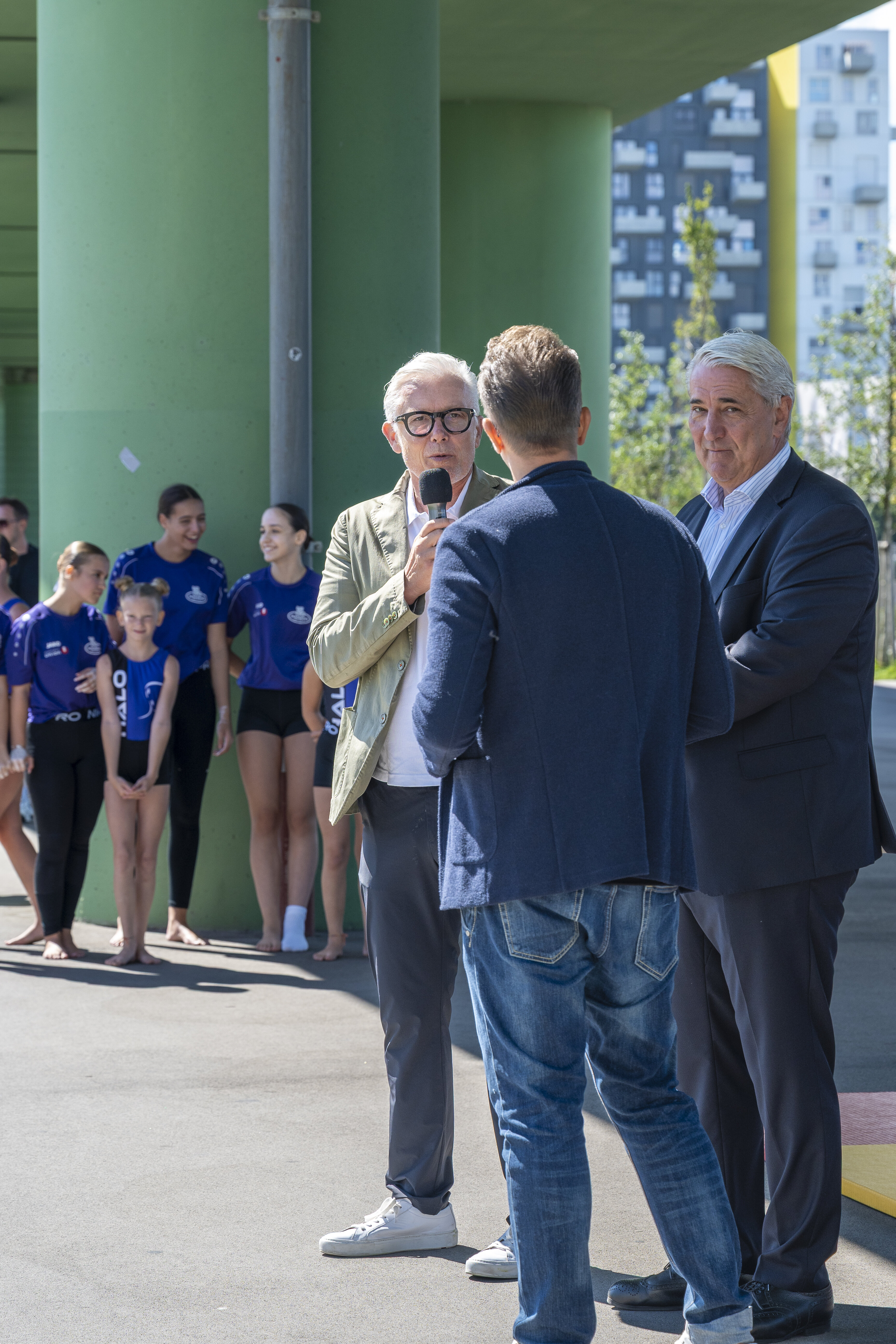 Robert Grüneis spricht in ein Mikrofon und steht in der Gruppe mit Bezirksvorsteher Ernst Nevrivy, im Hintergrund ist eine Gruppe Mädchen zu sehen, die in Sportoutfits gekleidet sind. 