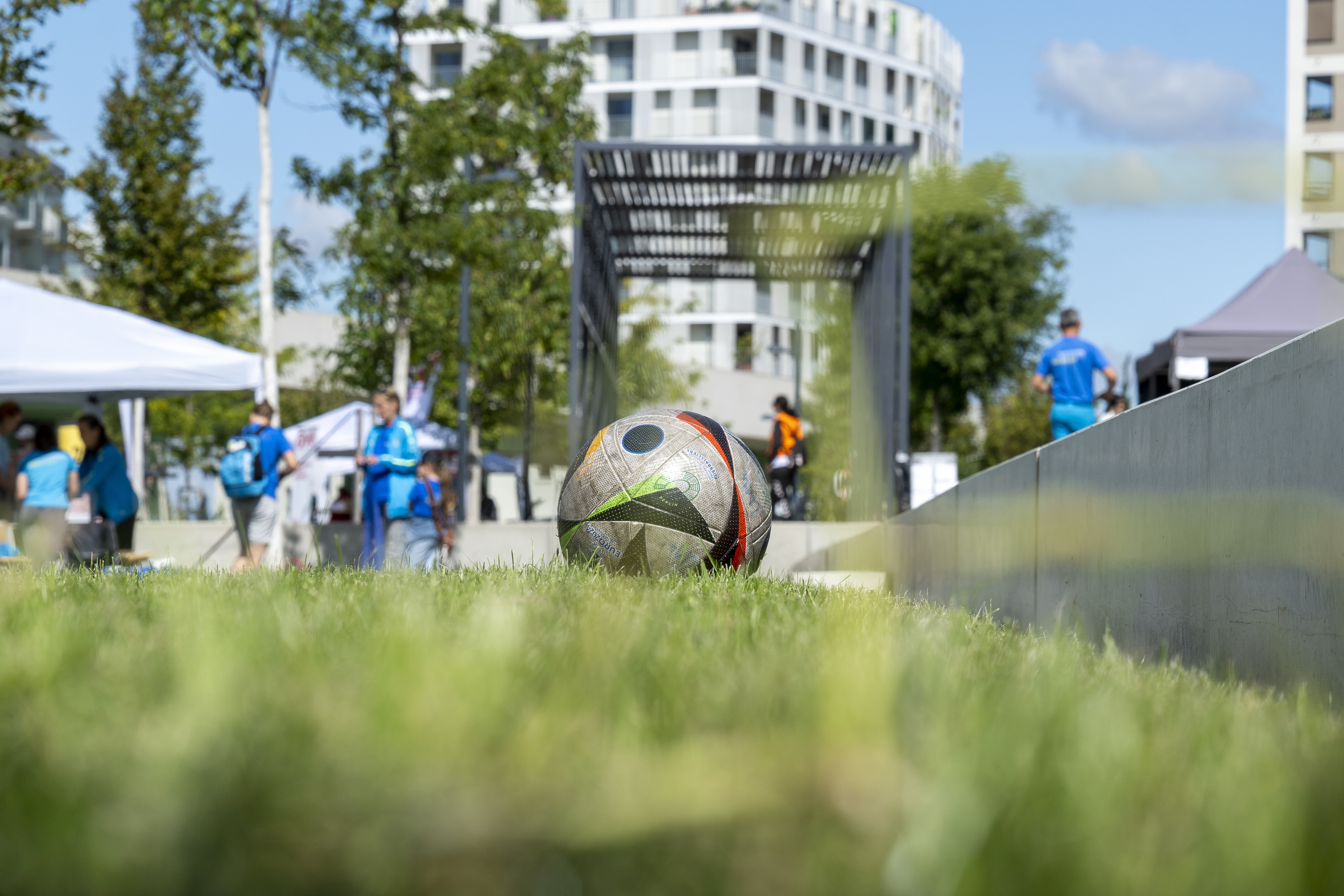 Ein Fußball liegt im Gras, im Hintergrund sind Menschen und Häuser zu sehen. 