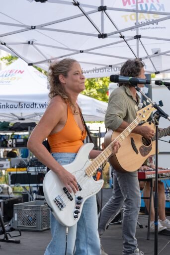 Frau mit orangefarbenem T-Shirt und einer Gitarre steht auf einer Bühne mit Mikrofon, im Hintergrund ist ein Mann mit Gitarre zu sehen