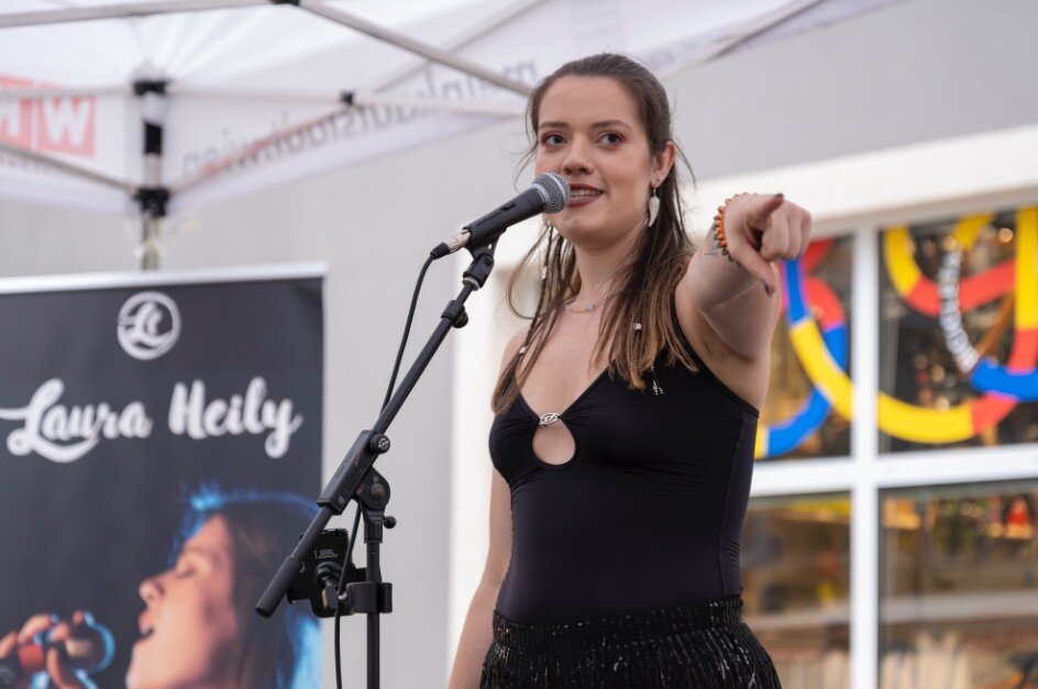 Sängerin  mit dunklen Haaren und schwarzem Trägerleiberl steht auf einer Bühne in einem Zelt und zeigt nach vorne. 