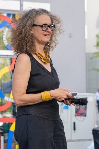 Frau mit Locken und schwarz gekleidet hält ein Mikrofon in der Hand und steht auf einer Bühne auf der Straße 