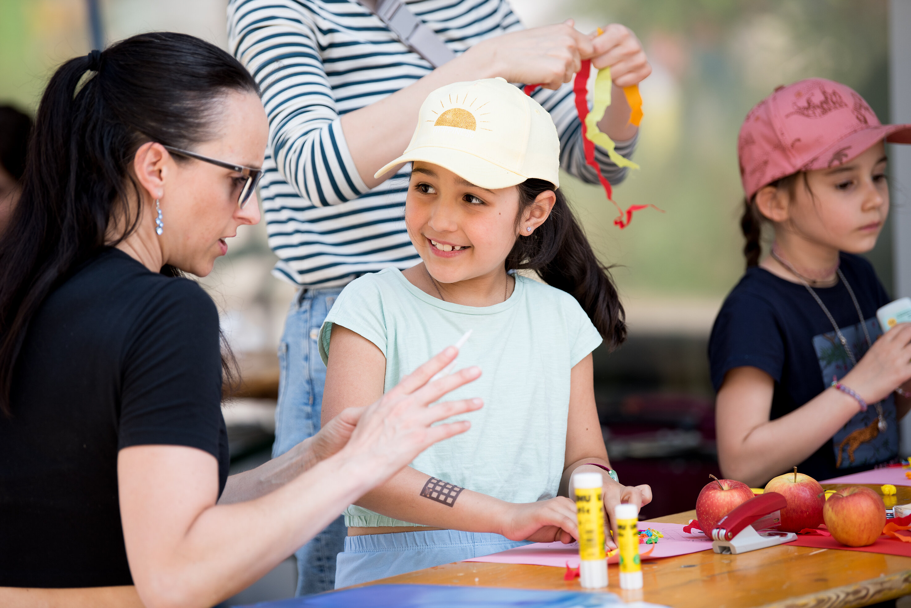 Eine Frau mit zwei Kindern sitzt an einer Biergarnitur und bastelt, die Kinder sind geschminkt und tragen Käppis