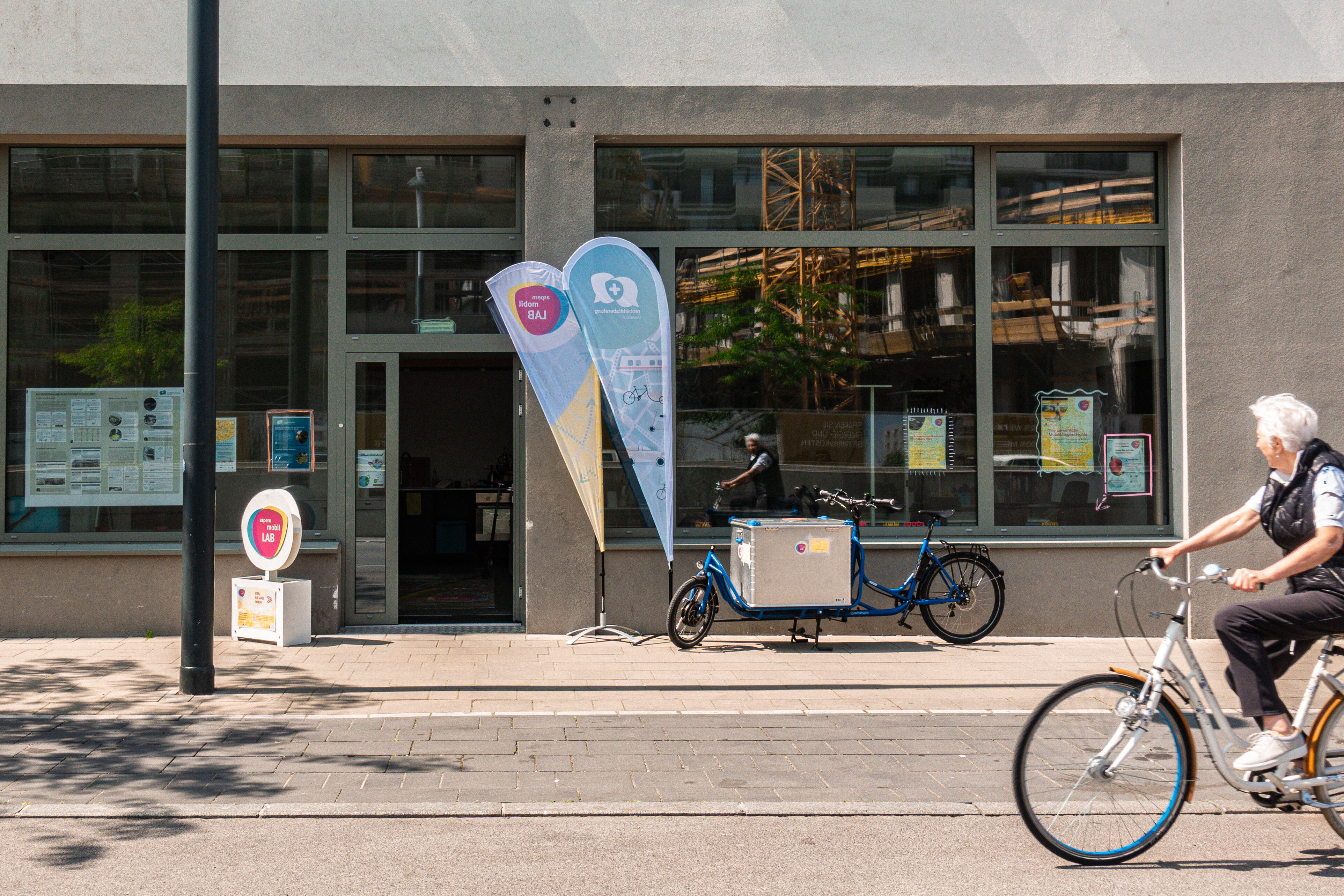 Ältere Frau auf einem Fahrrad fährt an einem Geschäftslokal von aspern.mobil. LAB vorbei