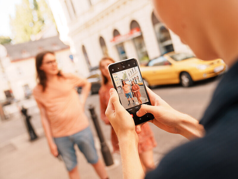 Eine Person macht ein Bild von zwei Frauen, es ist nur der Bildschirm des Handys und die Hände der fotografierenden Person scharf gestellt, der Rest ist verschwommen 