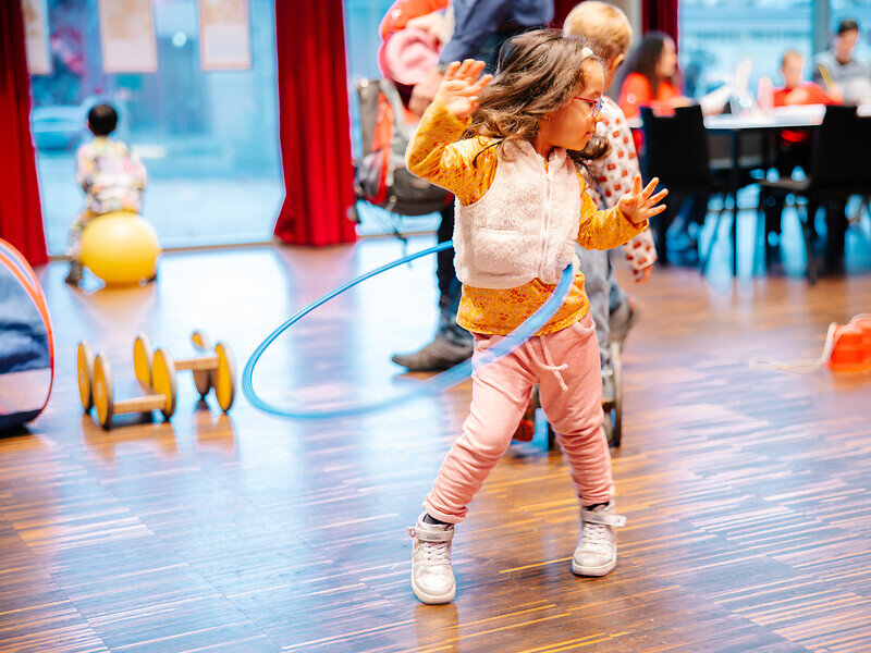 Ein Kind tanzt mit einem Hula-Hoop-Reifen, im Hintergrund sind andere Kinder zu erkennen, die sich verschieden bewegen