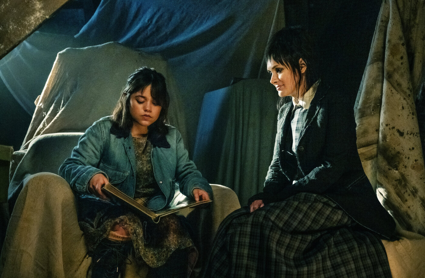 Bildausschnit aus dem Film Beetlejuice Beetlejuice, ein Kind sitzt mit einem offenen Buch auf dem Schoß auf einem Sack, eine Frau sitzt daneben und sieht zu