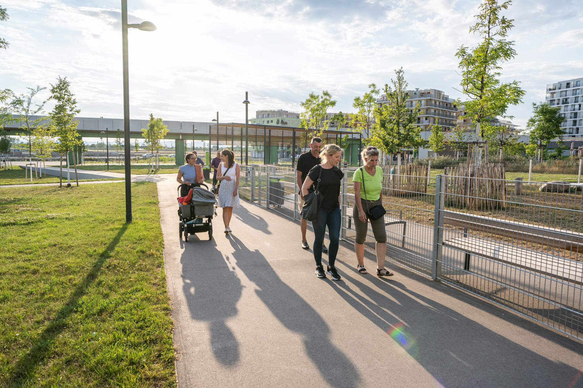 Eine Frau mit Kinderwagen, mehrere Frauen und 1 Mann spazieren und unterhalten sich dabei, es ist eine U-Bahntrasse und ein eingezäunter Bereich zu sehen
