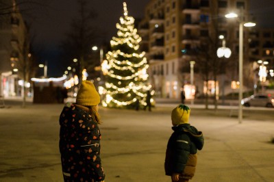 Es ist Abend. Zwei Kinder stehen am Hannah-Arendt-Platz und blicken in Richtung des beleuchteten Weihnachtsbaums.
