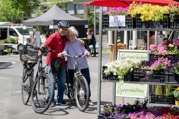 ein älteres Pärchen mit Fahrrädern
                                    umarmen einander neben einem Blumenstand