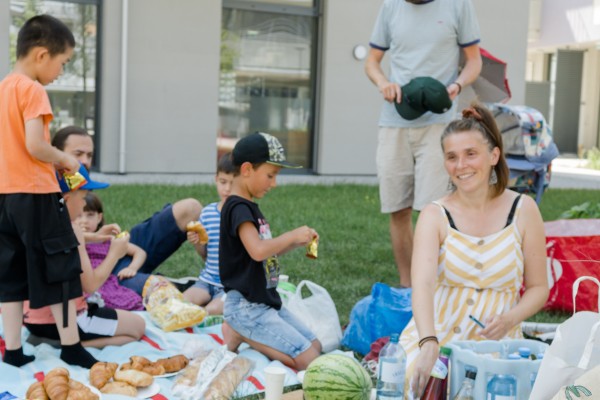 Erwachsene und Kinder sitzen auf einer Picknickdecke und man sieht Esse, wie eine Wassermelone, Chips, Humus und Brot