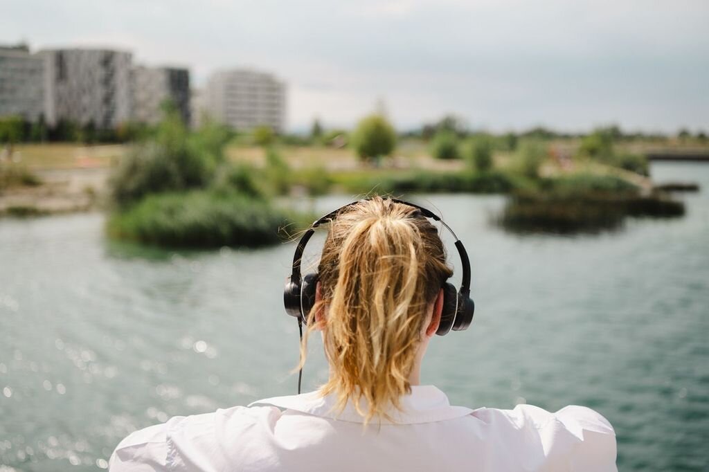 Eine Person mit blondem Pferdeschwanz und schwarzen Kopfhörern blickt von einem Ufer über das Wasser auf moderne Wohngebäude und Grünflächen in der Seestadt.