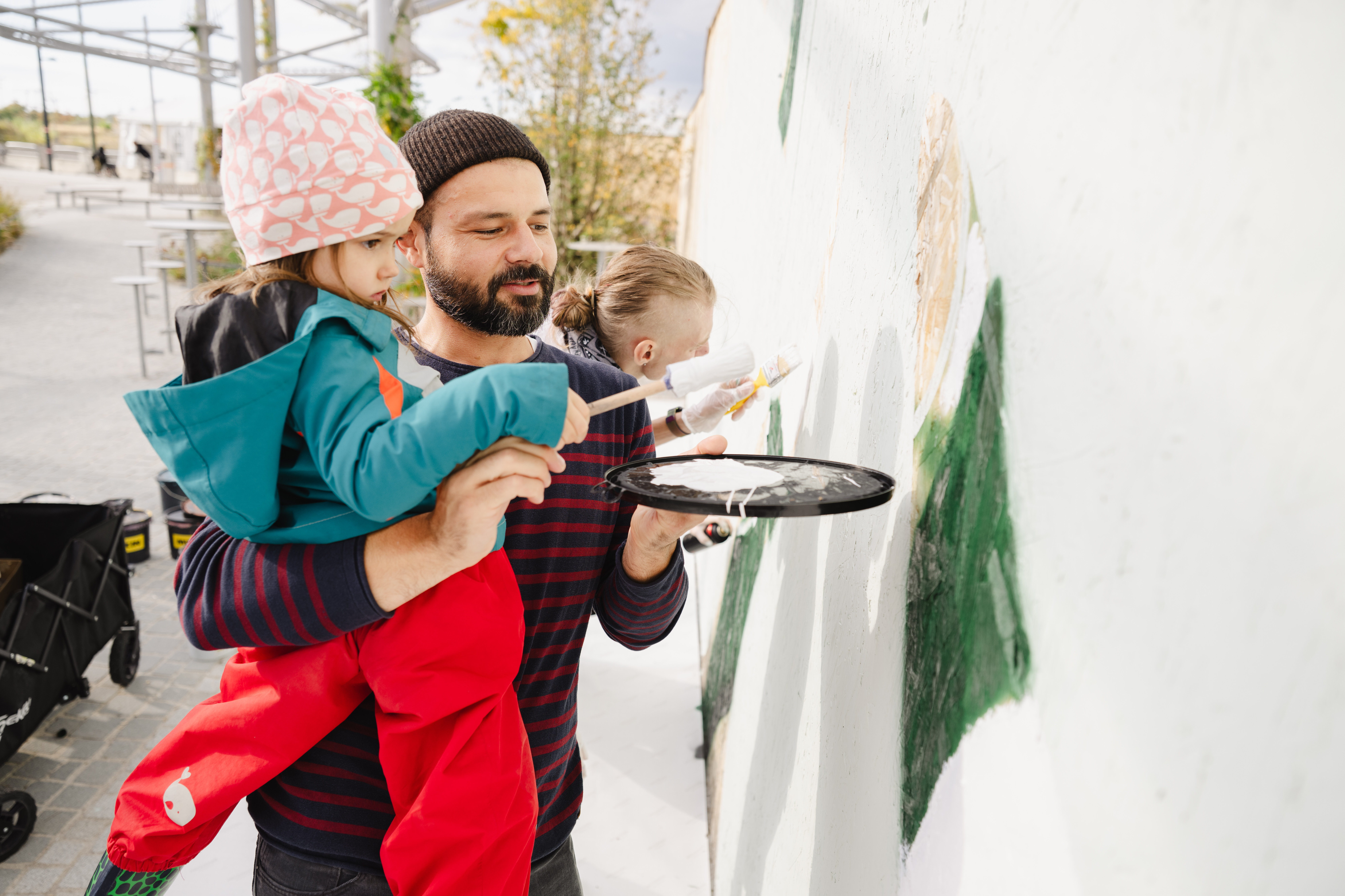 Ein Mann hält ein Kind im Arm, während beide gemeinsam eine Wand bemalen. Der Mann trägt eine Mütze und gestreiftes Oberteil, das Kind eine bunte Jacke und rote Hose. Im Hintergrund sind weitere Personen und eine überdachte Fläche zu sehen.
