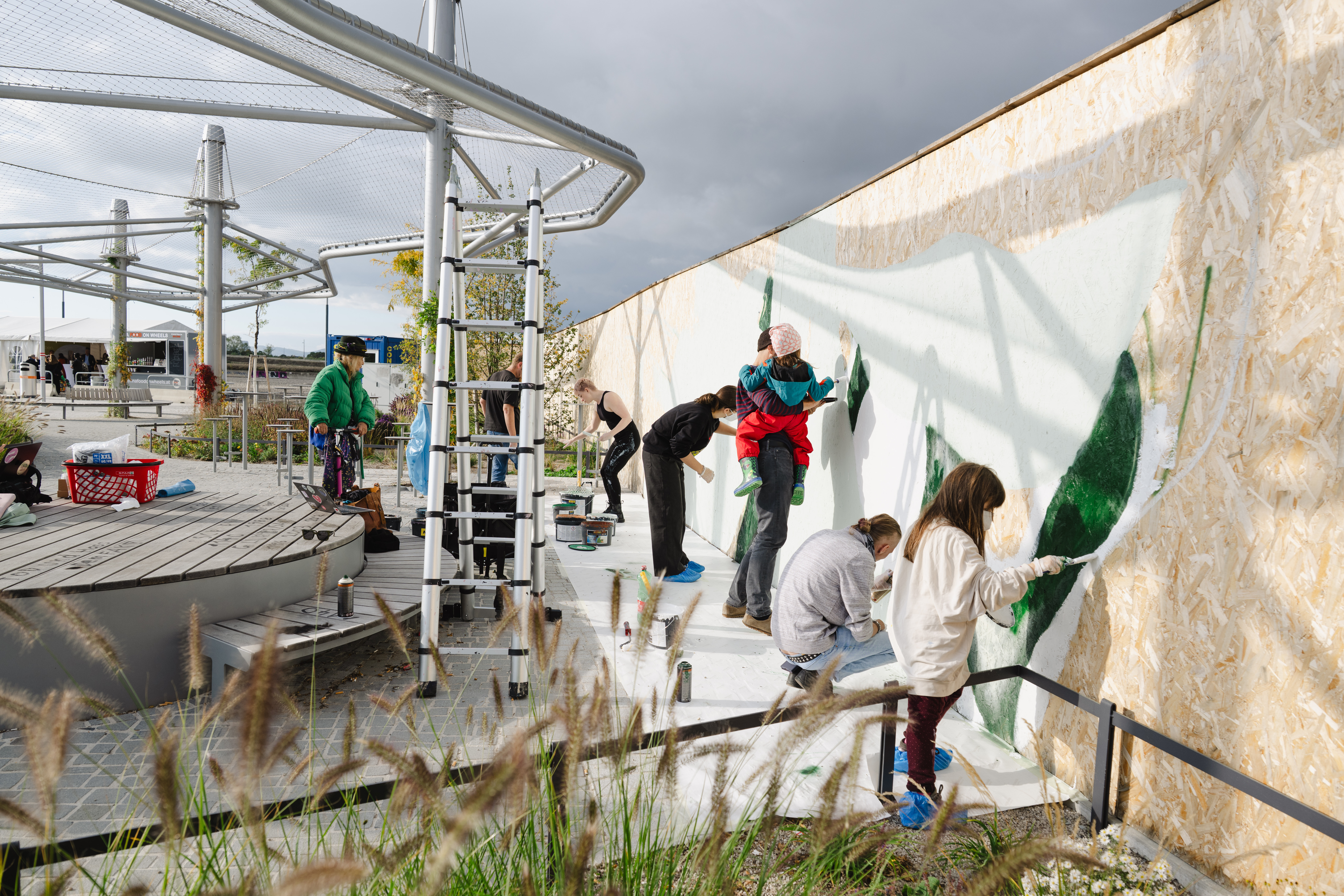 Mehrere Personen malen gemeinsam an einer großen Wand im Freien. Einige tragen Schutzkleidung und verwenden Pinsel und Farbrollen, um grüne Flächen auf eine helle Wand aufzutragen. Im Vordergrund sind Pflanzen, im Hintergrund eine moderne Überdachung aus Metall.