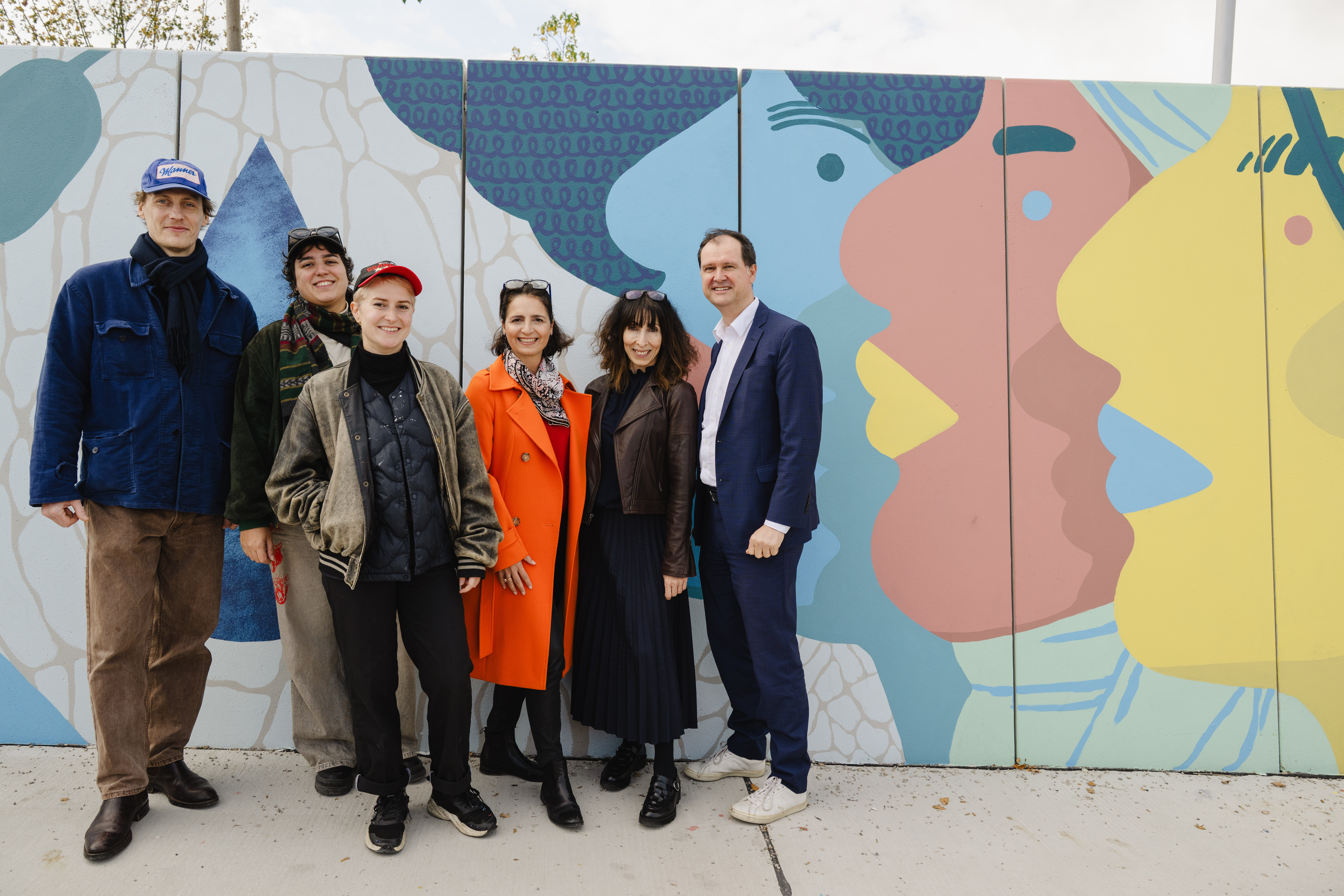 Eine Gruppe von sechs Personen steht lächelnd vor einer bunt bemalten Wand mit abstrakten Gesichtern in Blau, Rosa und Gelb.