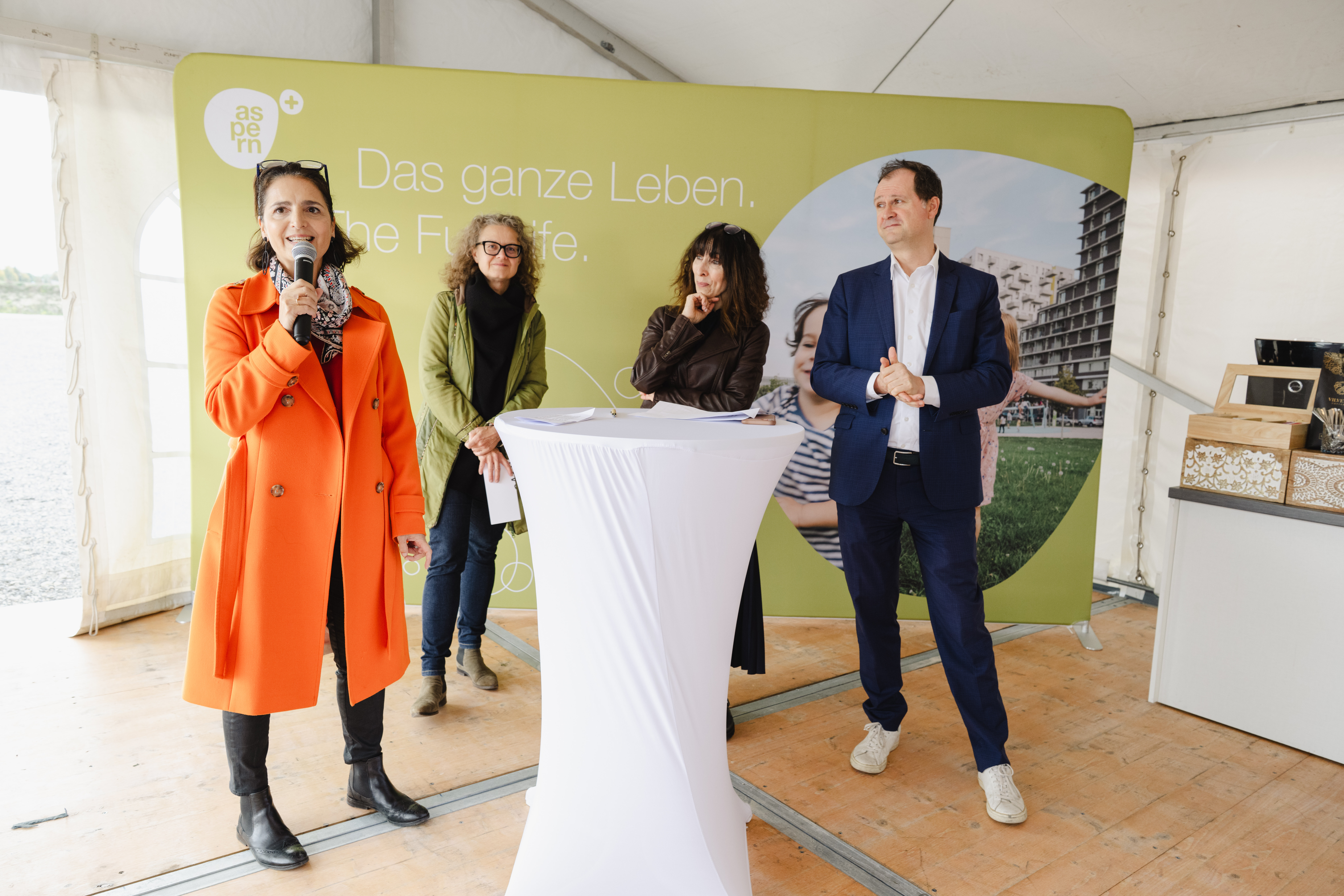 Vier Personen stehen unter einem Zelt vor einer grünen Wand mit der Aufschrift „Das ganze Leben. The Full Life.“. Eine Frau im orangefarbenen Mantel spricht in ein Mikrofon, während die anderen zuhören.