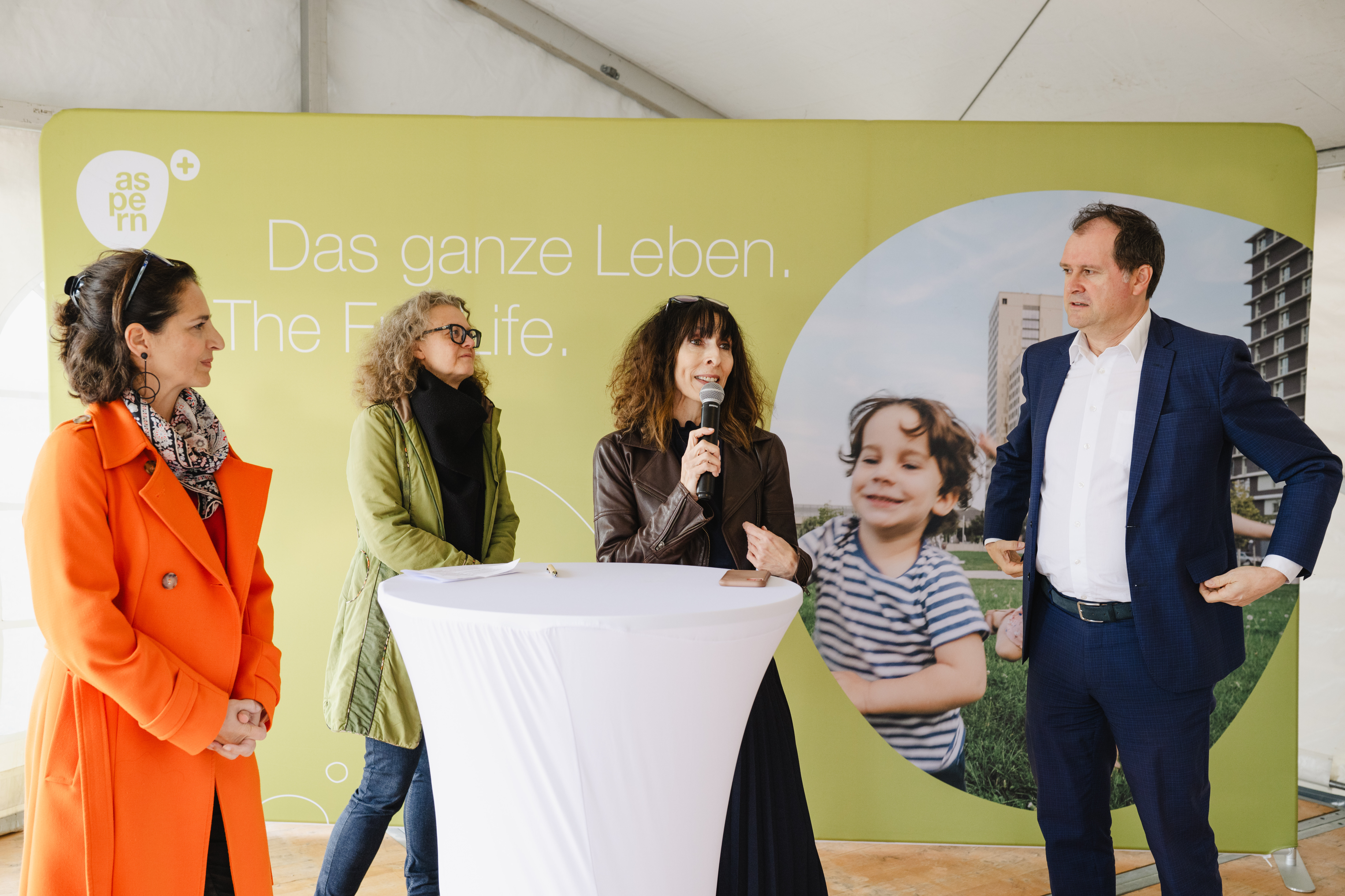 Vier Personen stehen an einem weißen Stehtisch unter einem Zelt. Eine Frau mit dunklen Haaren spricht in ein Mikrofon, während die anderen zuhören. Im Hintergrund ist ein grünes Banner mit dem Text „Das ganze Leben. The Full Life.“