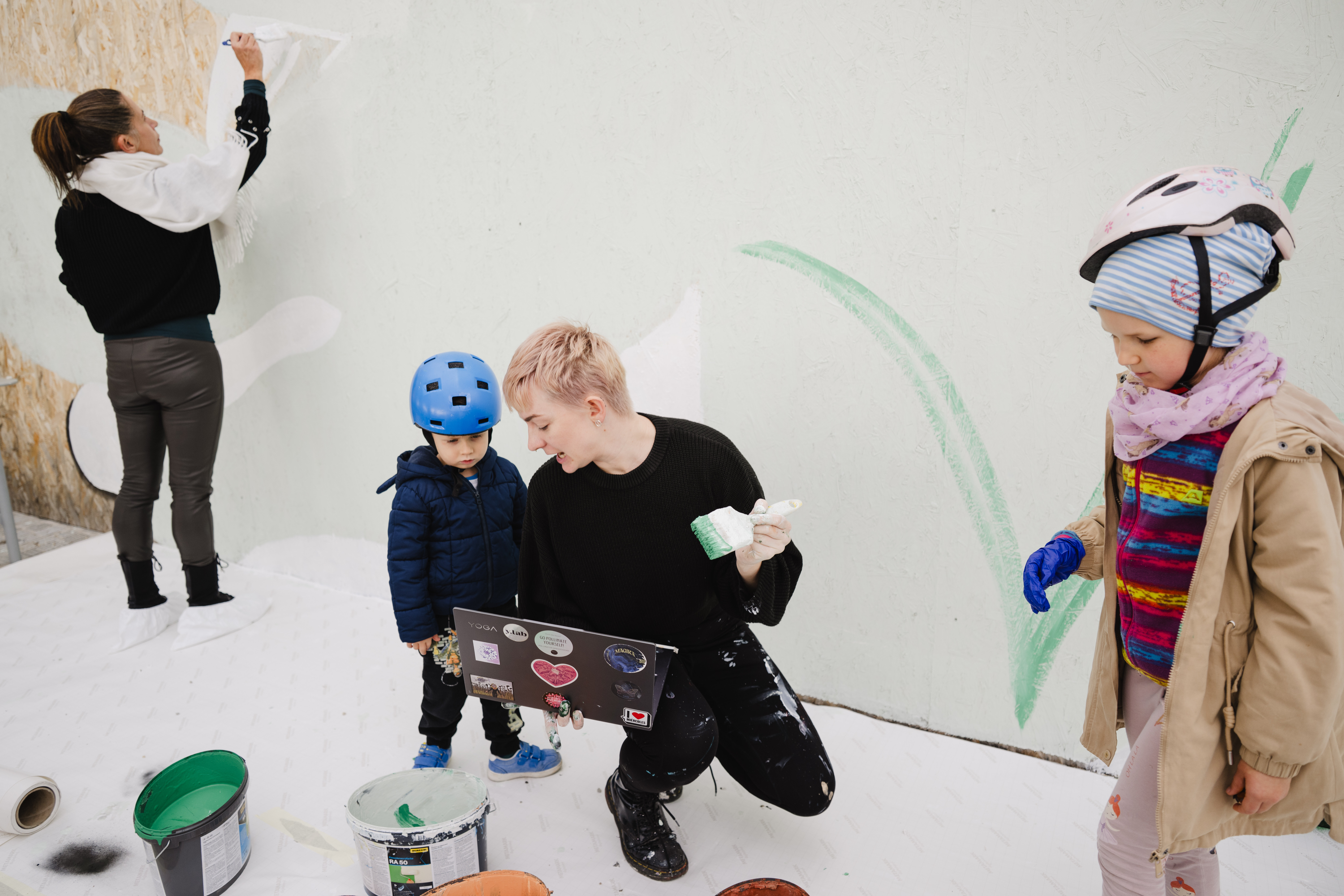 Eine Frau malt mit einem kleinen Kind an einer Wand. Neben ihnen stehen Farbdosen und zwei weitere Kinder mit Helmen, die ebenfalls am Malprojekt teilnehmen.